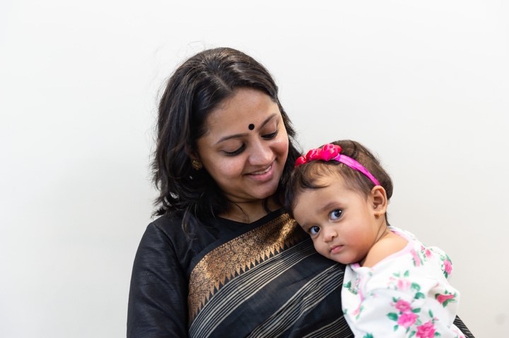 Rashmi smiles as her daughter rests her head on her shoulder looking at the camera
