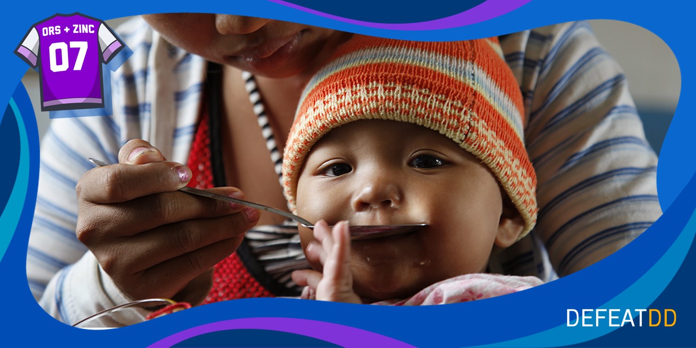 A mother feeds her baby with a spoon. The baby, wearing a knitted hat, looks at the camera. The image has a blue border with a purple shirt icon labeled "ORS + ZINC 07" and the word "DEFEATDD" at the bottom.