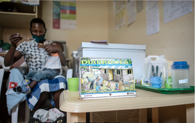 A healthcare setting with a woman feeding a baby. In the foreground, a booklet titled "Diarrhoea" is on a table, surrounded by medical supplies. The room has educational posters on the walls.