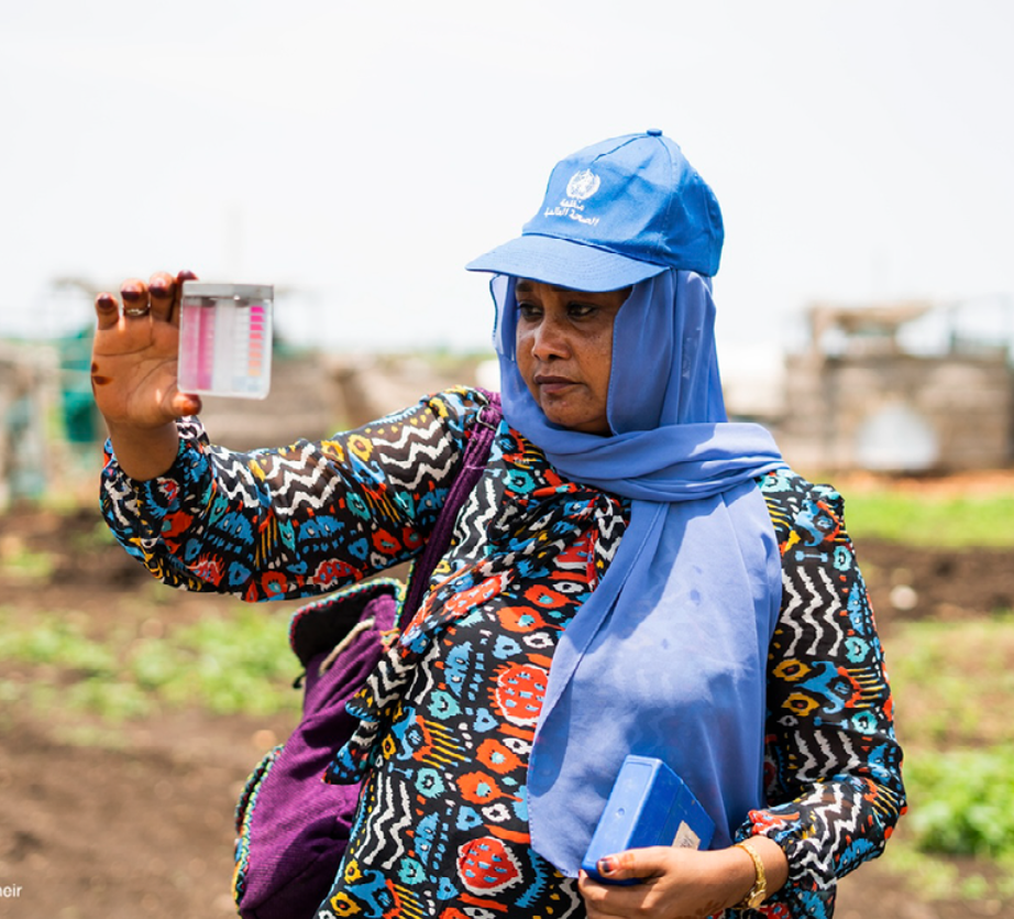 A person wearing a colorful shirt and a blue cap, holding a water testing kit in one hand and observing it closely. They are standing outdoors in a field with greenery around, under a clear sky.