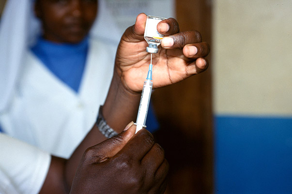 A person is drawing liquid from a vial into a syringe. Another person in the background is wearing a white uniform with a blue shirt underneath. The background has a wall with blue and beige sections.