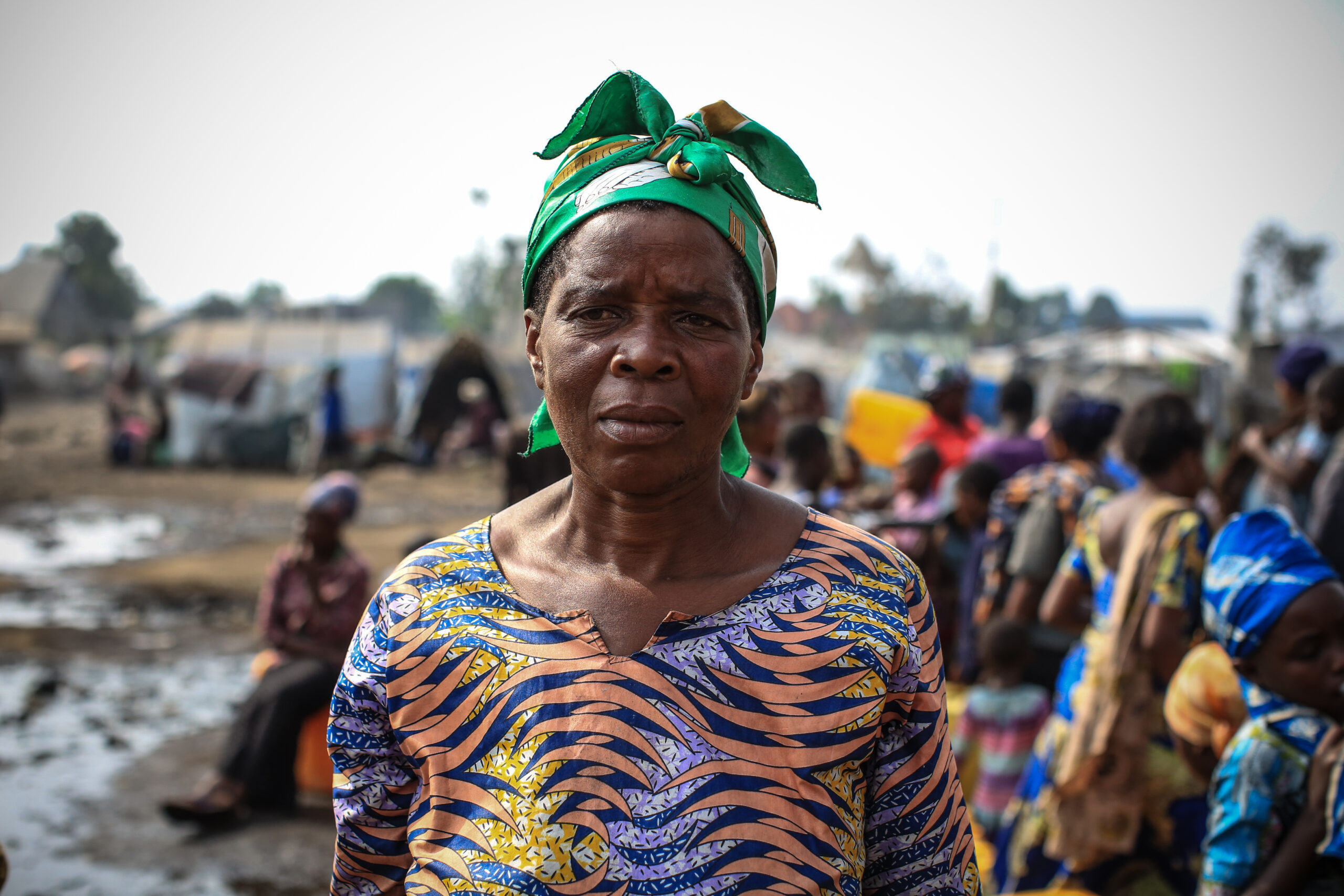 A woman wearing a green headscarf and a colorful patterned dress stands outdoors. People and makeshift tents are seen in the blurry background, suggesting a crowded, possibly refugee or camp setting. The atmosphere appears busy and the ground looks muddy.