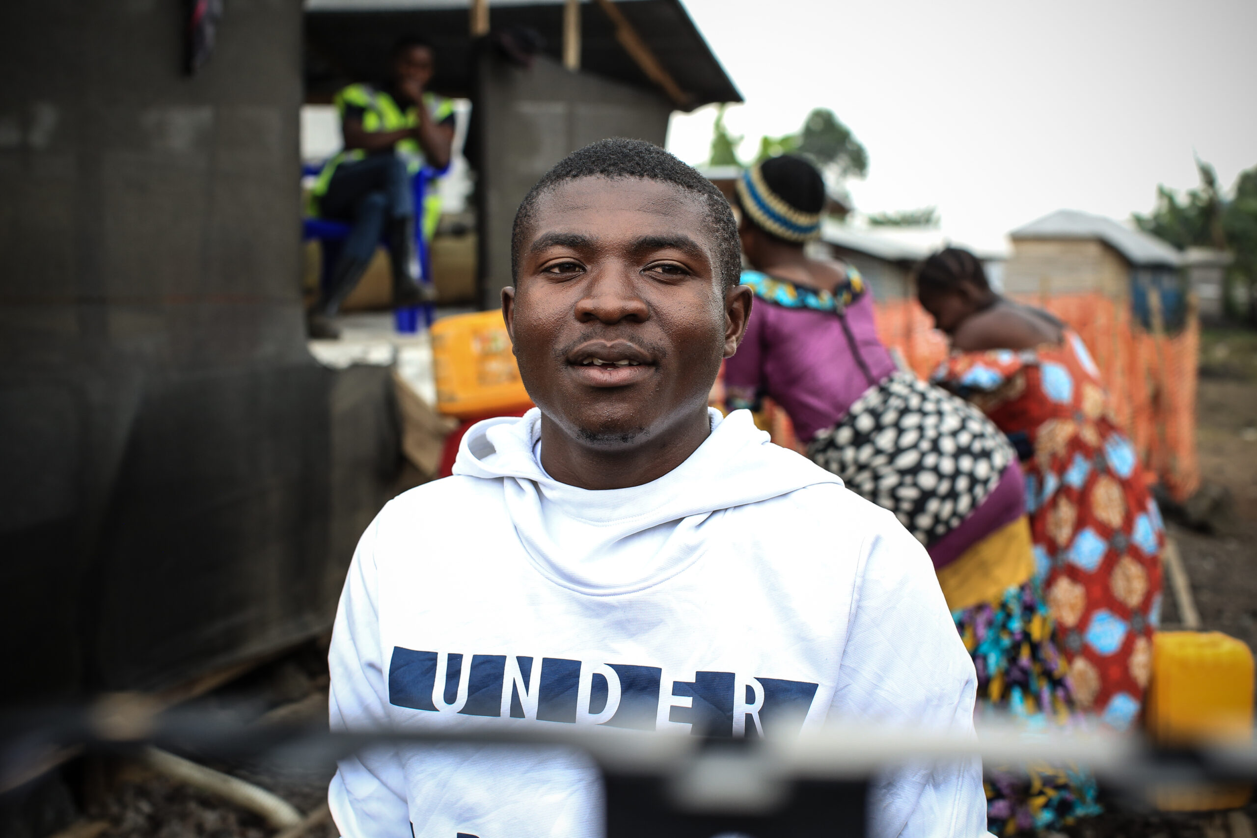 A person wearing a white hoodie stands outdoors with an open-mouthed expression. In the background, people in colorful clothing are engaged in various activities near a modest building. A person in a green uniform sits on a chair.