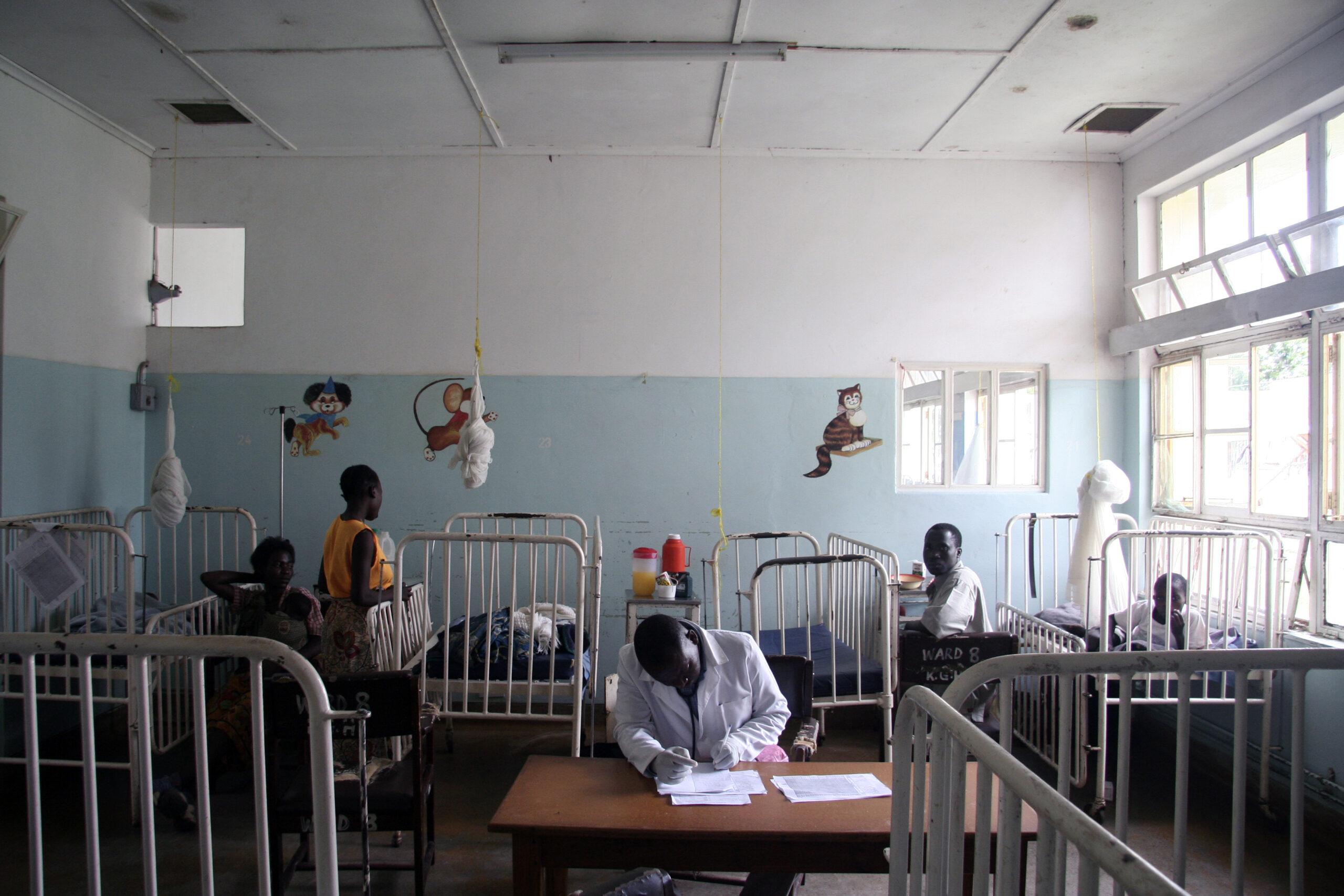 A healthcare worker in a white coat sits at a desk, writing in a notebook in a room with multiple cribs and beds. A few adults and children are present, with colorful illustrations of animals on the walls. The room is lit by natural light from a window.