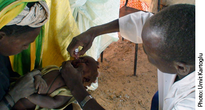 A healthcare worker administers an oral vaccine to a baby held by a woman. The scene takes place indoors with colorful fabric in the background.