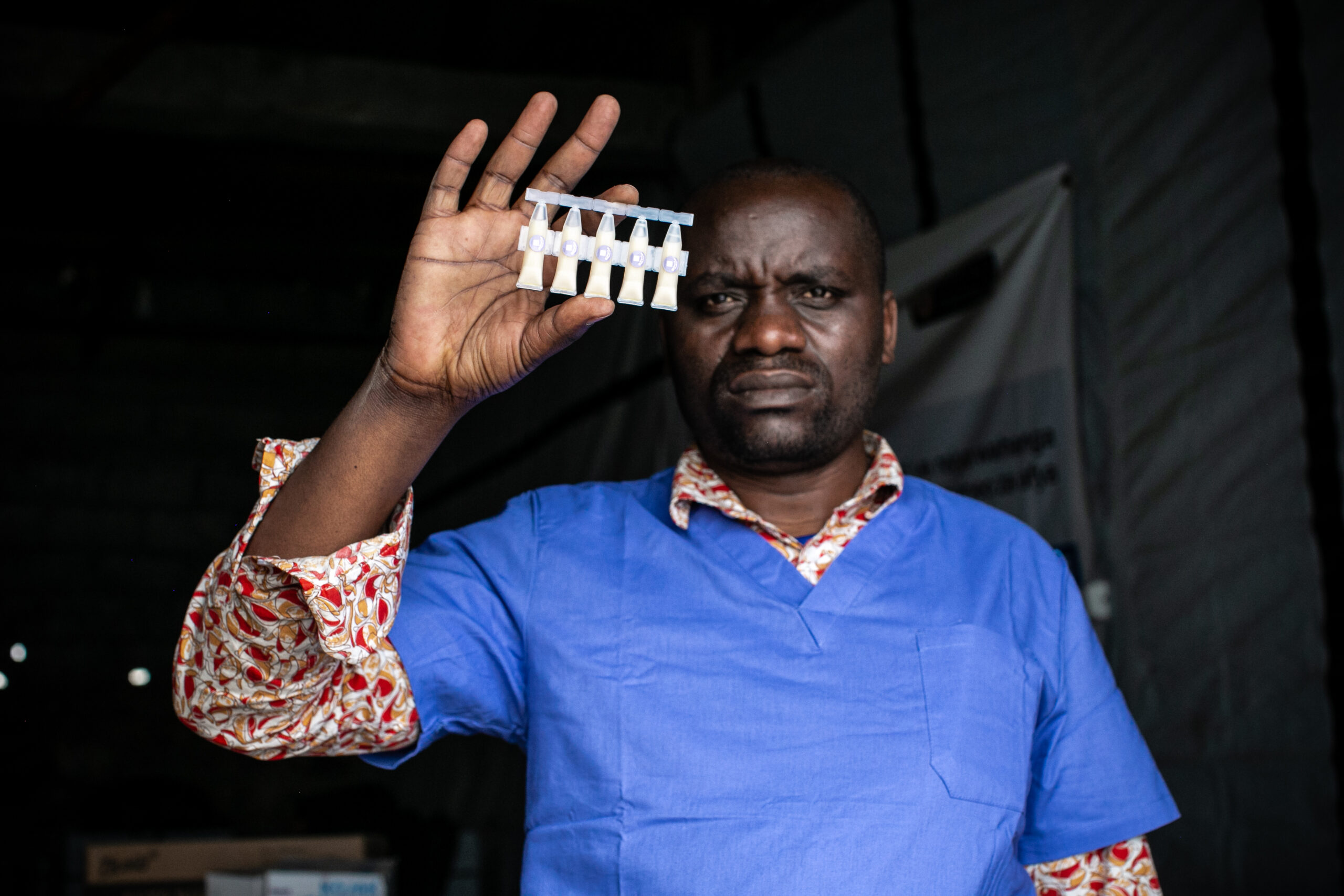 A person wearing a blue coat and a colorful patterned shirt holds a strip of white medical vials up to the camera, showing them prominently. The individual has a serious expression and stands indoors with a dark background.