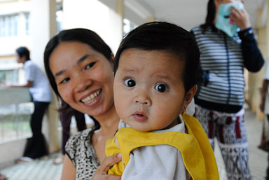 A smiling woman holds a baby who is looking at the camera with a curious expression. The baby is wearing a yellow garment. In the background, another person is wearing a mask and using their phone.