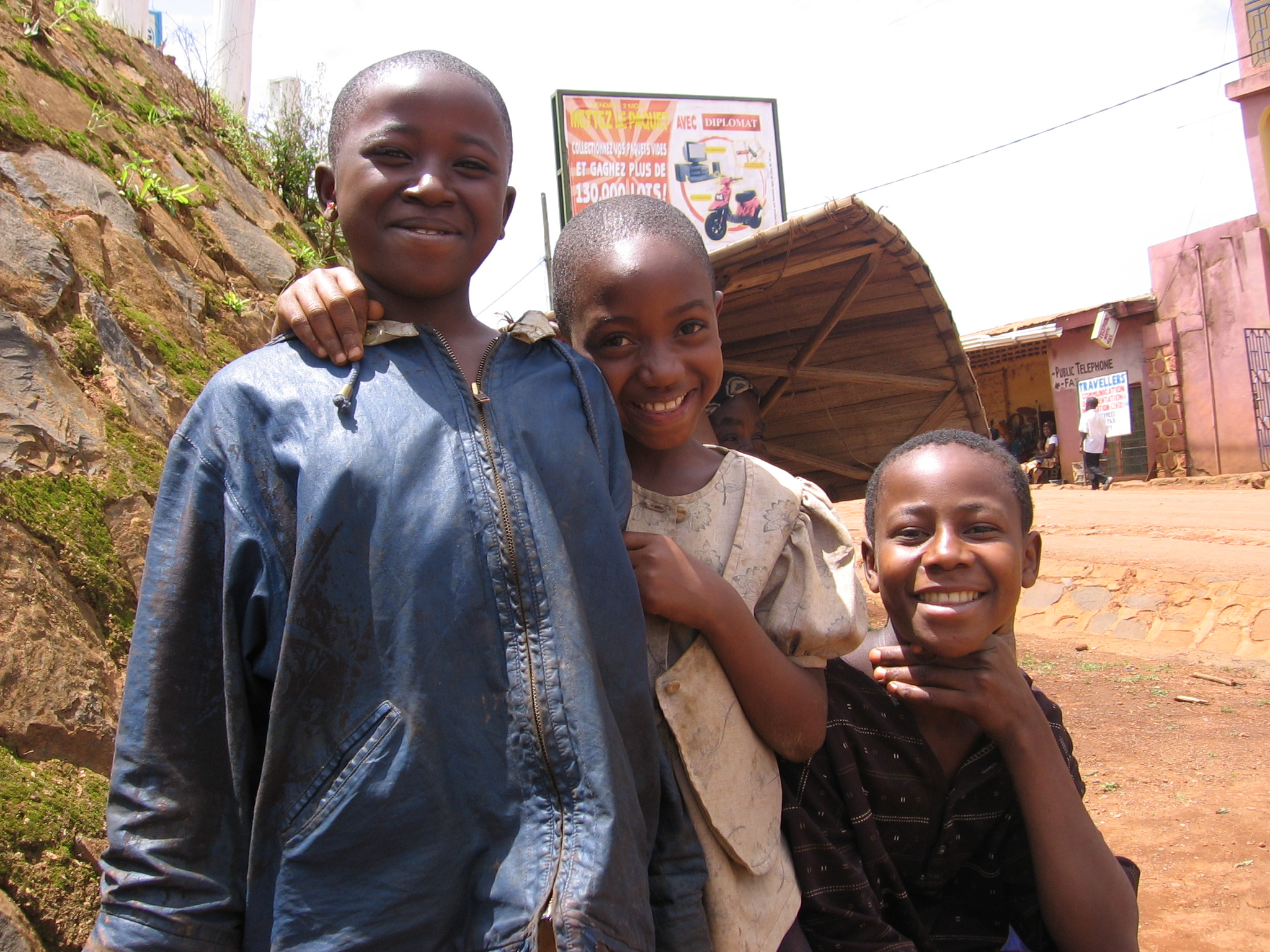 Three smiling children pose for the camera on a sunny day. One child on the left wears a blue jacket, the middle child has an arm around the first child and wears a beige shirt, and the third child on the right wears a black shirt. They stand on a dirt road.