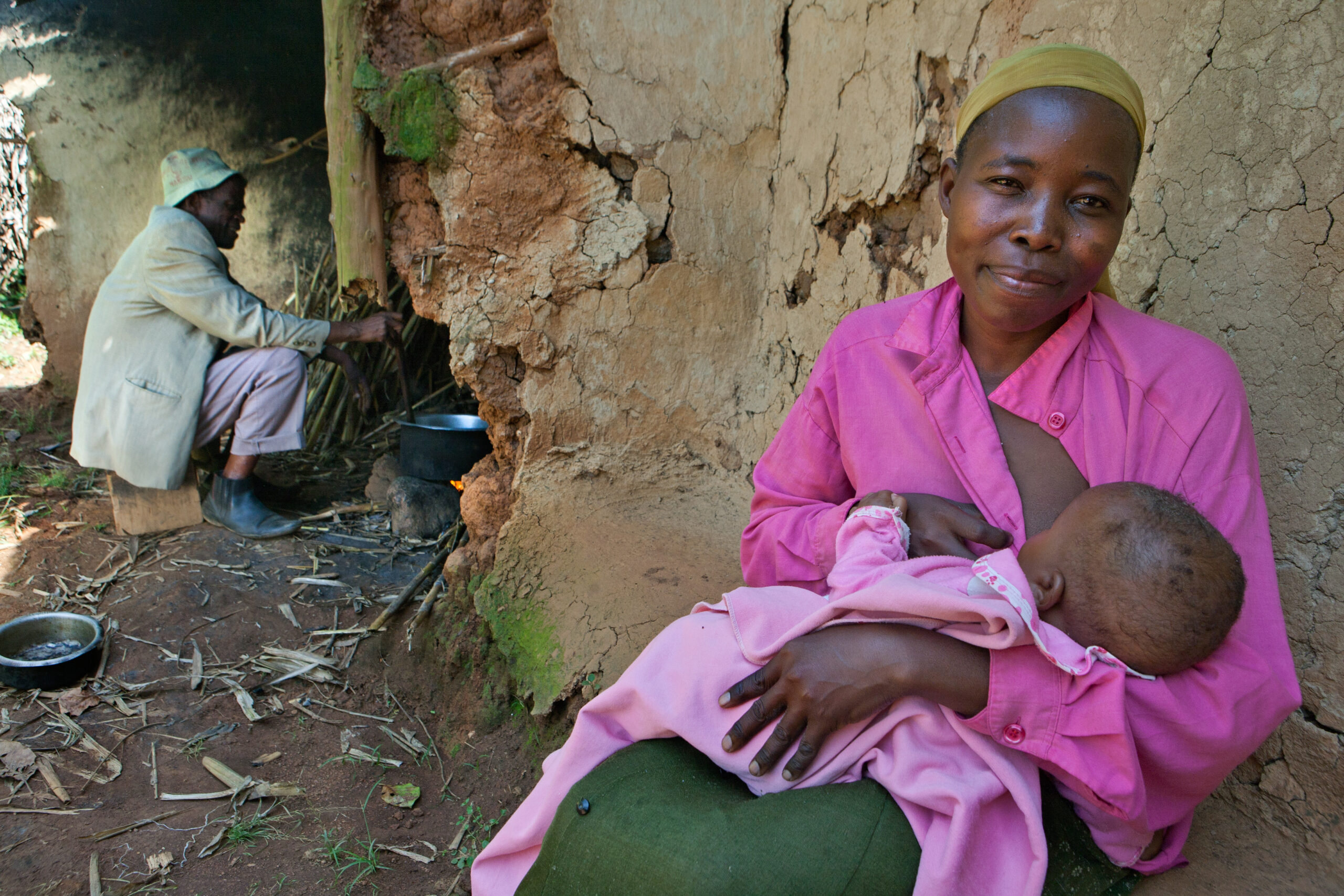 A woman with short hair and a headscarf, dressed in a pink shirt, is sitting against a rustic wall, breastfeeding an infant in pink clothing. In the background, a man in a hat is crouched by a cooking pot near a small fire. The setting appears rural.