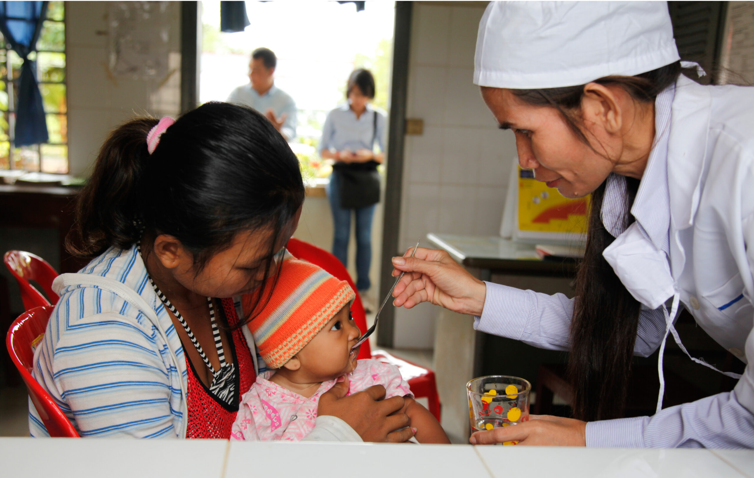 A healthcare worker in a white uniform and cap administers medicine to a baby held by a woman. The baby wears an orange and white hat. The mother and baby are seated on red chairs, with other people seen in the blurred background of the room.