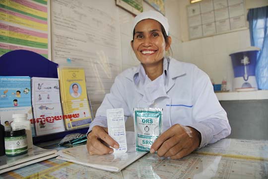 A smiling healthcare worker in a white uniform and headscarf is seated at a desk. They hold up packets of Oral Rehydration Salts (ORS). Various health posters and educational materials are displayed on the wall in the background.