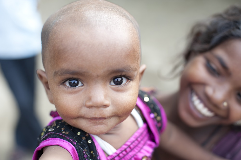 A close-up of a baby with large dark eyes, wearing a purple outfit, looking directly into the camera. In the background, a woman with a bright smile, slightly out of focus. The image conveys warmth and affection.