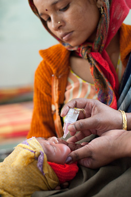 A woman wearing a headscarf watches as an adult administers medicine or drops to a baby wrapped in a colorful blanket. The baby is held by the adult's hands, with a focus on the medicine being given.