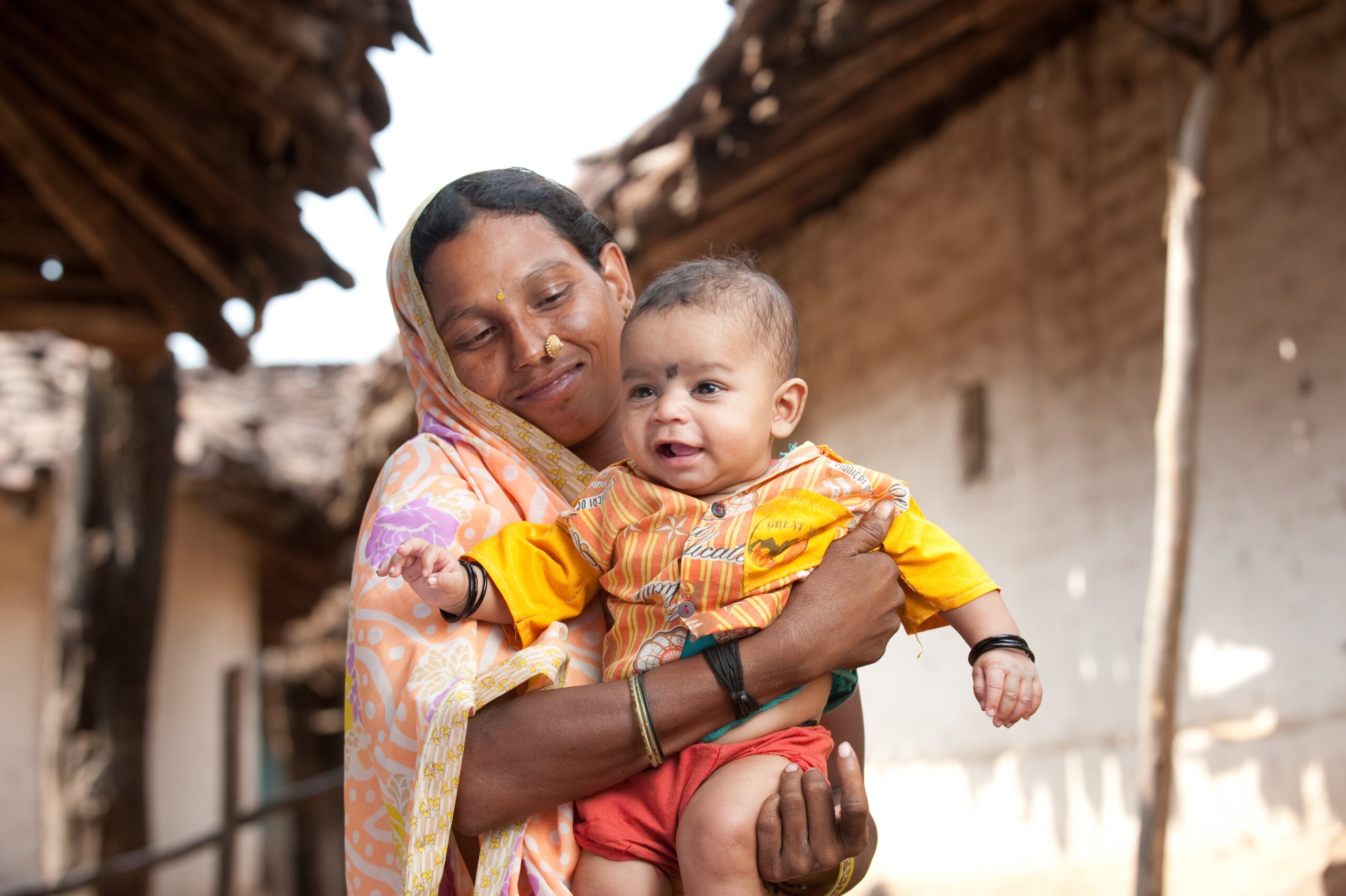 A smiling woman dressed in a colorful sari holds a happy baby wearing an orange outfit. They are outside near rustic buildings with thatched roofs. The baby appears joyful and the woman looks at the baby lovingly. The background suggests a rural setting.