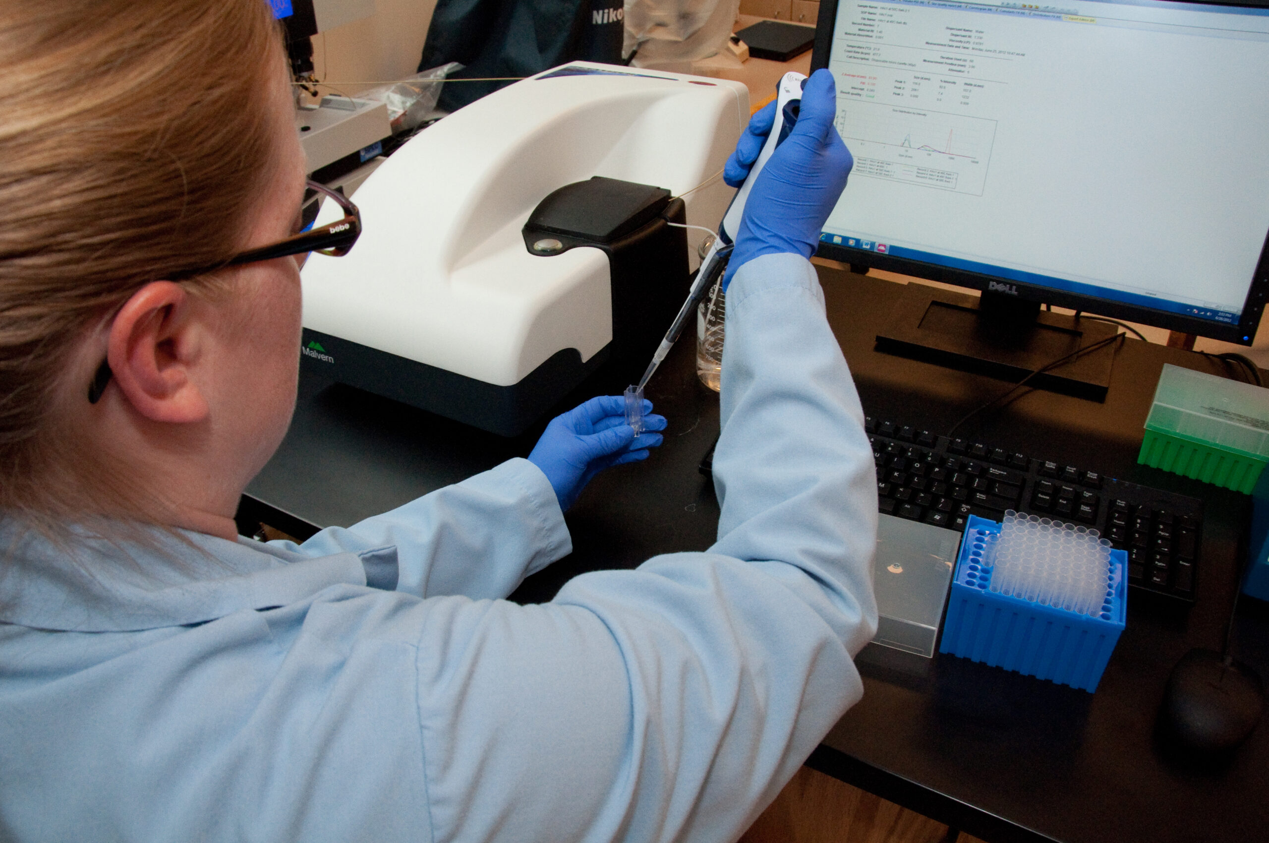 A person wearing a lab coat and blue gloves is holding a pipette and working at a desk with scientific equipment. A computer screen displays data, and a tray of test tubes sits nearby.