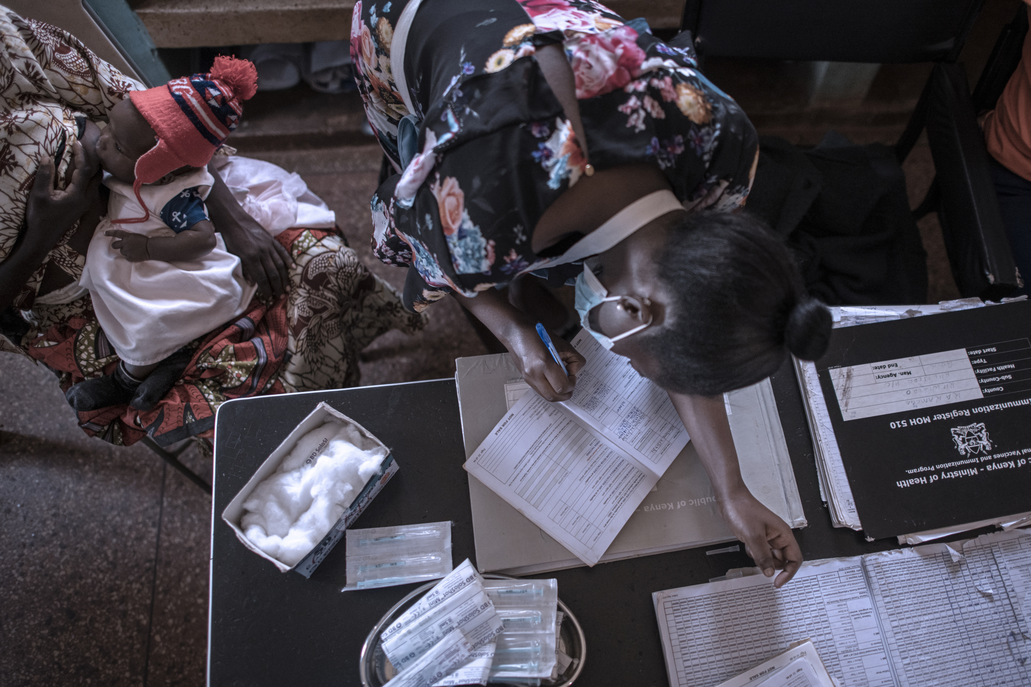 A woman leans over a table filling out paperwork. Nearby, a person holds a baby wearing a red hat. Various medical supplies are scattered on the table. The scene suggests a clinic or medical setting.