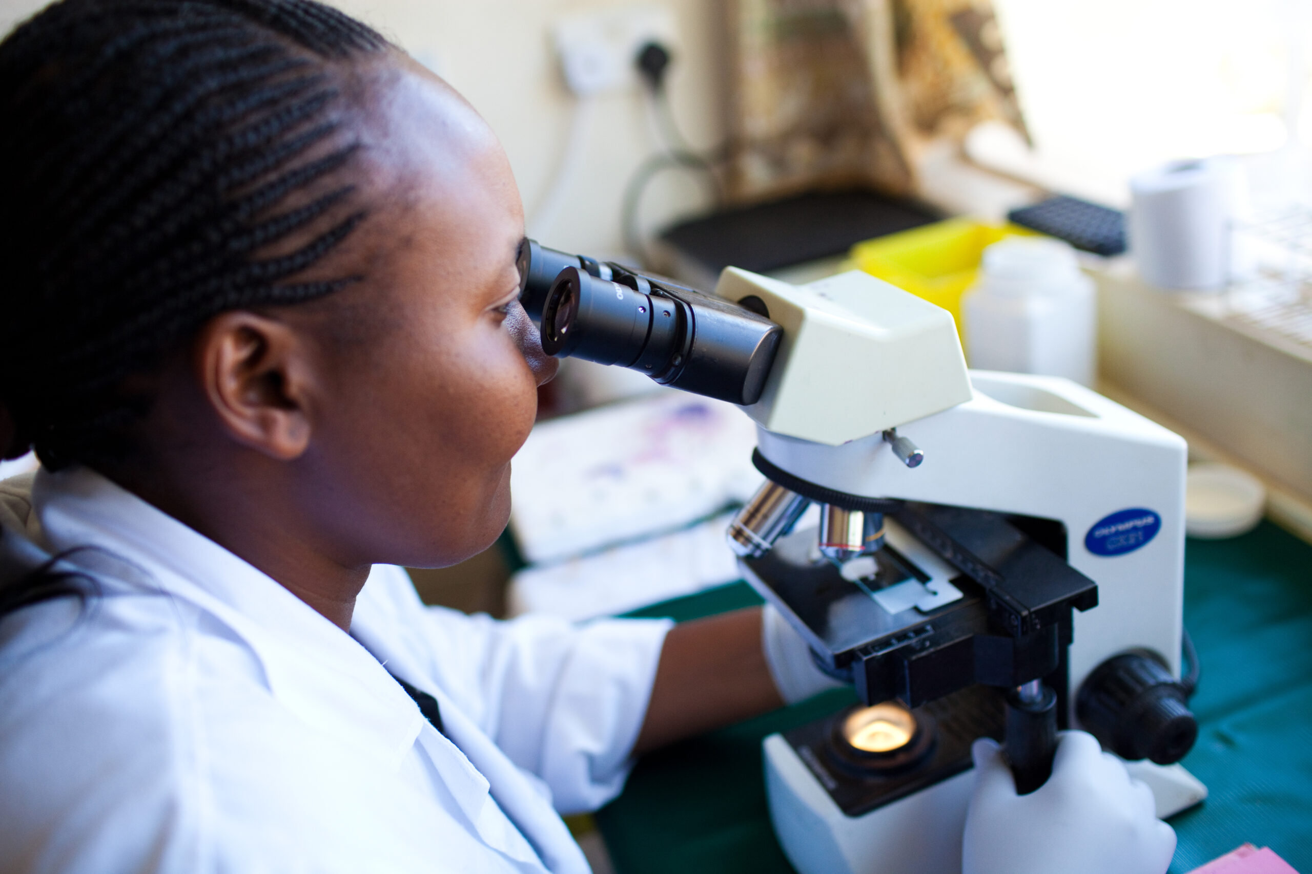 A person wearing a white lab coat and gloves is looking through a microscope in a laboratory setting. There are various lab equipment and containers on the table, and a window with natural light in the background.