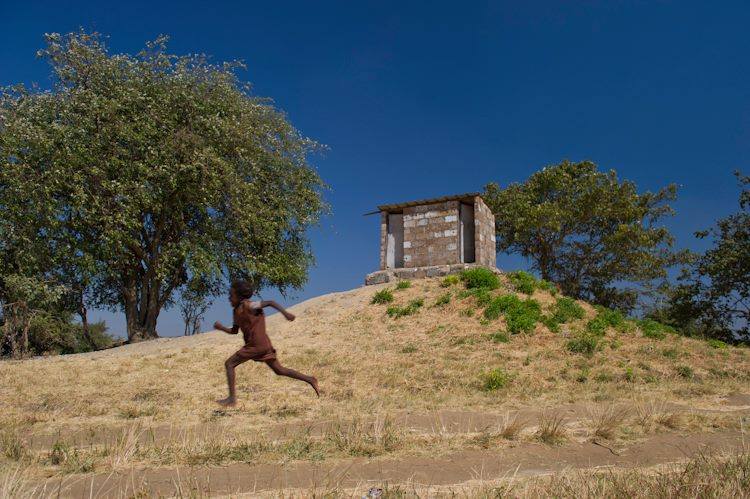 A person runs up a grassy hill with bare feet, wearing a brown outfit. Trees and a small stone structure are in the background under a clear blue sky.