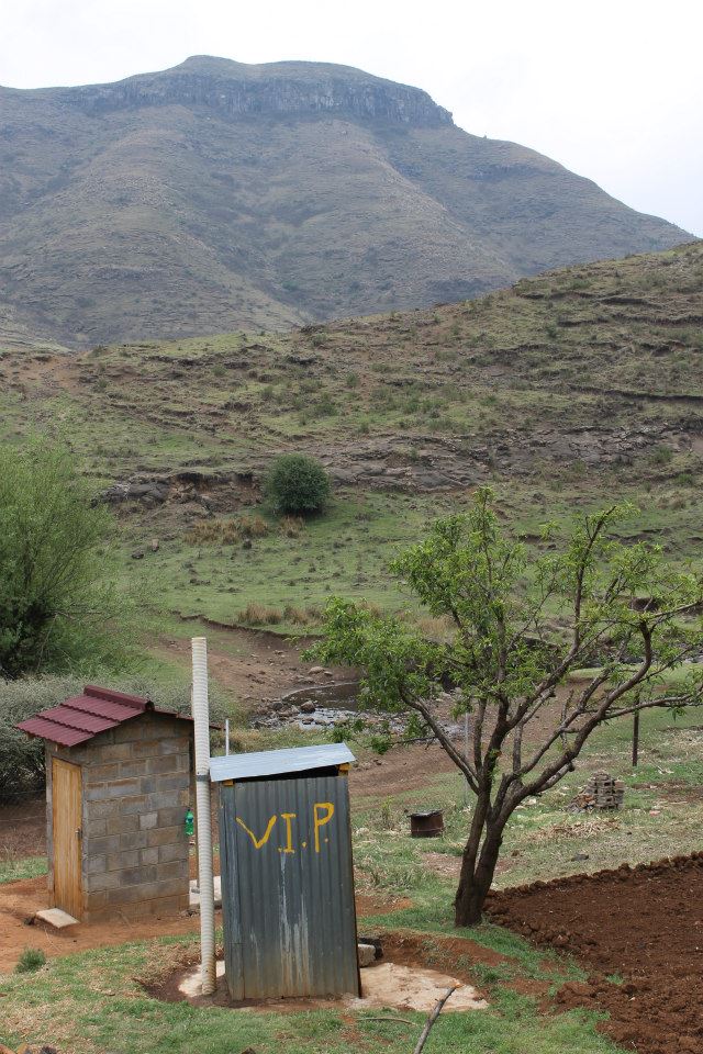 A rural scene with an outdoor pit latrine marked "V.I.P" in yellow paint. The latrine is next to a small brick structure with a red roof. The background shows grassy, hilly terrain with a tall mountain under a cloudy sky. A tree is also visible near the latrine.