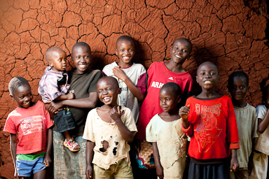 A group of happy children standing in front of a cracked earthen wall. They are smiling and embracing each other, with some wearing colorful t-shirts. The vibrant expressions capture a joyful moment.