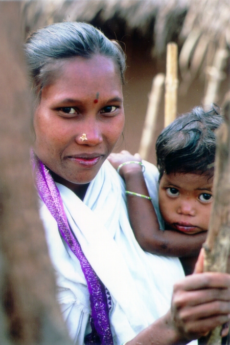 A woman with a bindi and nose ring in traditional attire stands holding a young child close to her. The child, wearing a bangle, clings to her shoulder, both looking directly at the camera with a thatched roof structure visible in the background.