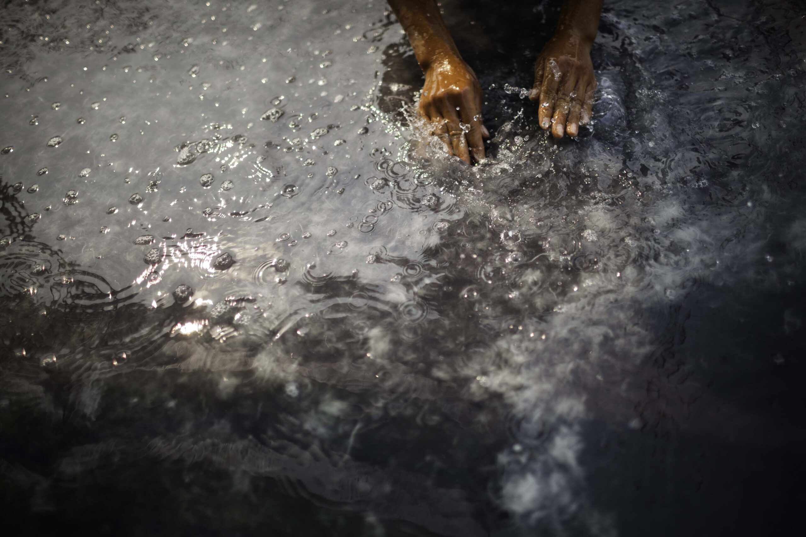 Hands reaching into a body of water, with ripples and bubbles visible on the surface. The lighting creates a silvery sheen on the water.