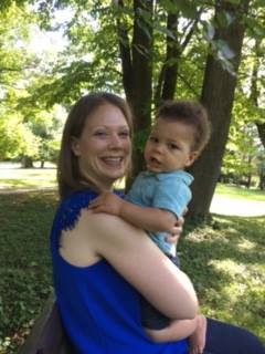 A woman with shoulder-length hair, wearing a blue sleeveless top, is sitting on a bench outdoors, holding a baby dressed in a light blue shirt. They are both smiling, surrounded by trees and greenery on a sunny day.