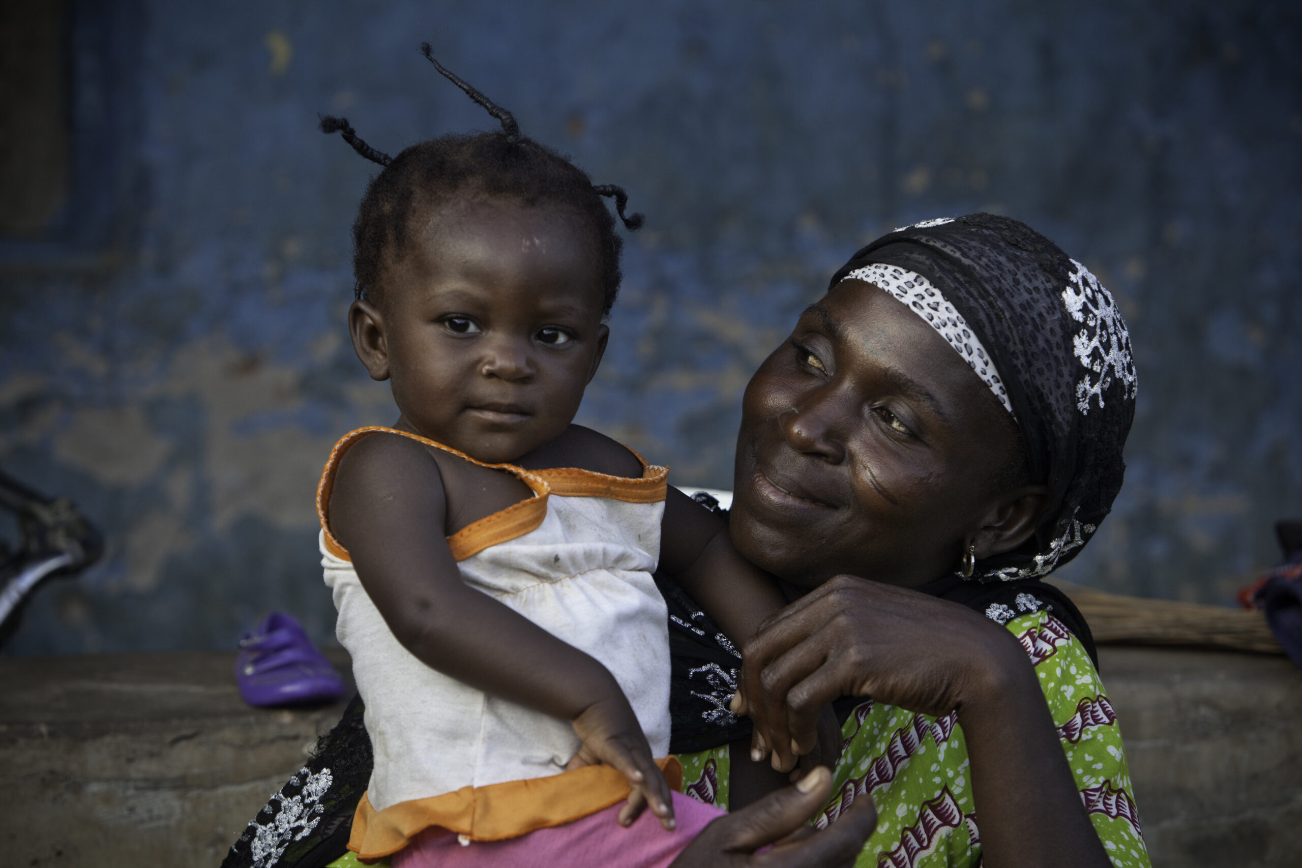 A woman wearing a patterned headscarf and dress lovingly holds a young child in her arms. The child is wearing a sleeveless top and gazing forward. They are sitting against a textured blue wall, sharing a tender moment.