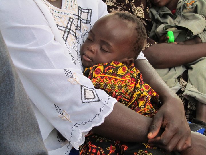 A small child is asleep, resting in the arms of an adult. The child is wearing a colorful patterned garment, while the adult's garment is white with black and gold embroidery. Other people are visible in the background.