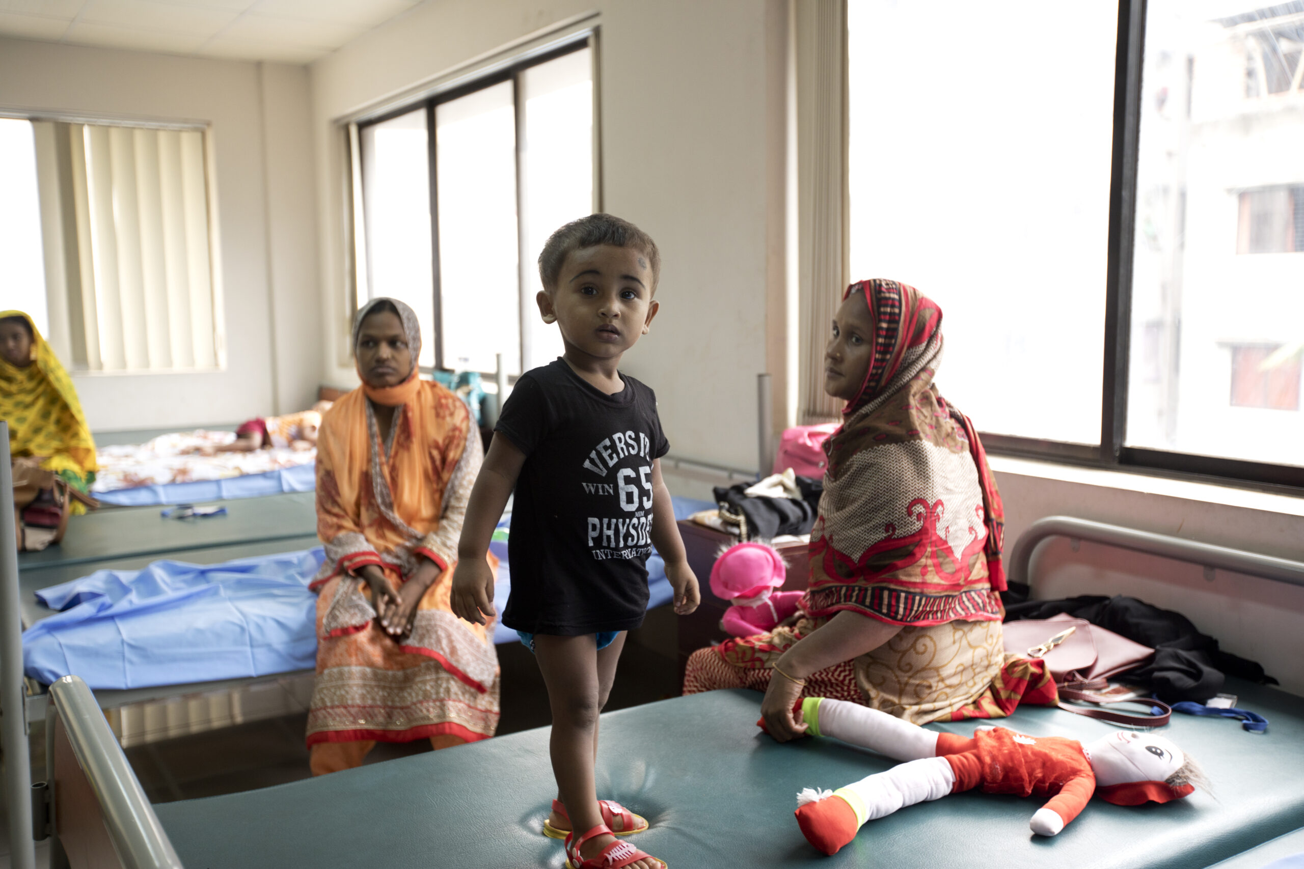 A young child stands on a hospital bed in a room with three women seated on other beds. The child is wearing a black shirt and shorts, and plush toys are scattered on the bed. Large windows let in natural light, highlighting the room's minimal furnishings.