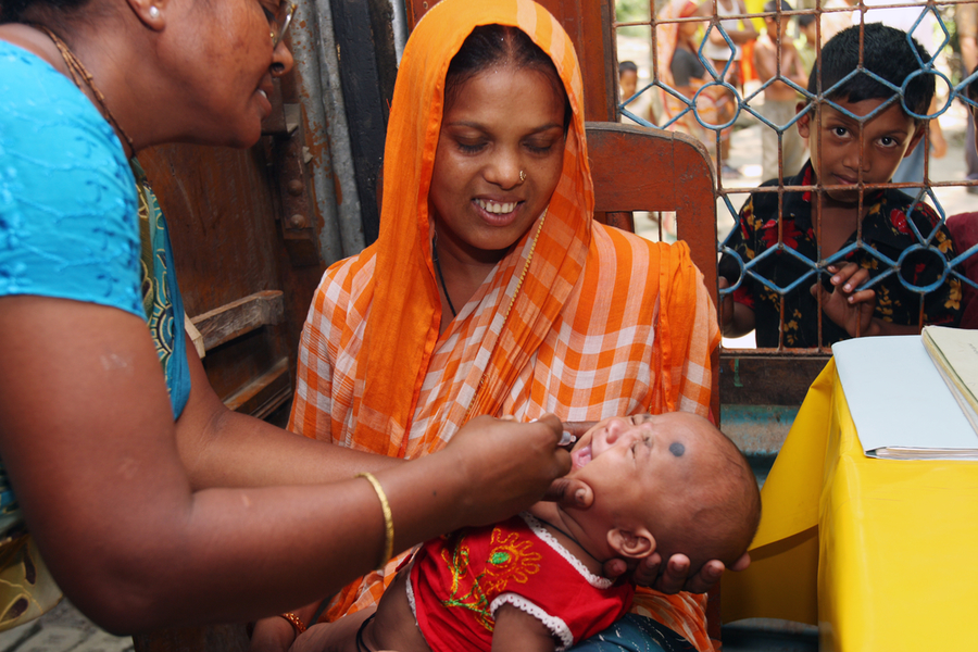 A woman in an orange and white checkered sari holds a baby while another woman administers oral medication to the baby. They are seated outdoors near a metal gate with a young boy observing from behind the gate. A yellow surface is partially visible.