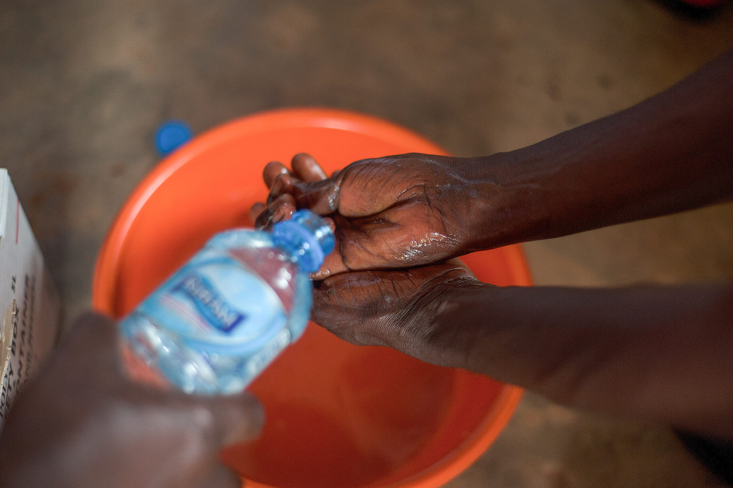 A close-up of hands being washed with water from a plastic bottle over an orange basin. The person's skin is dark, and the water is pouring onto their hands, creating a splashing effect. The background is slightly blurred.