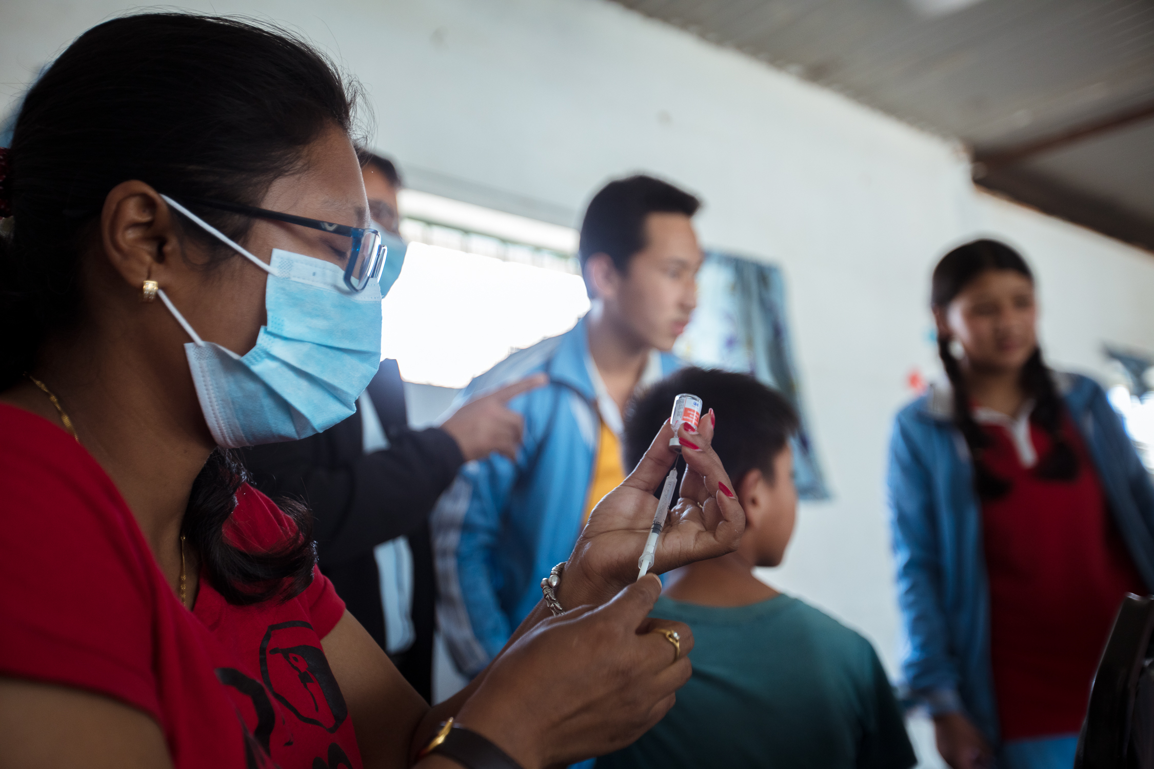 A healthcare worker wearing a mask prepares a syringe in the foreground. In the background, several people, including a child, wait in a bright room with blue curtains.