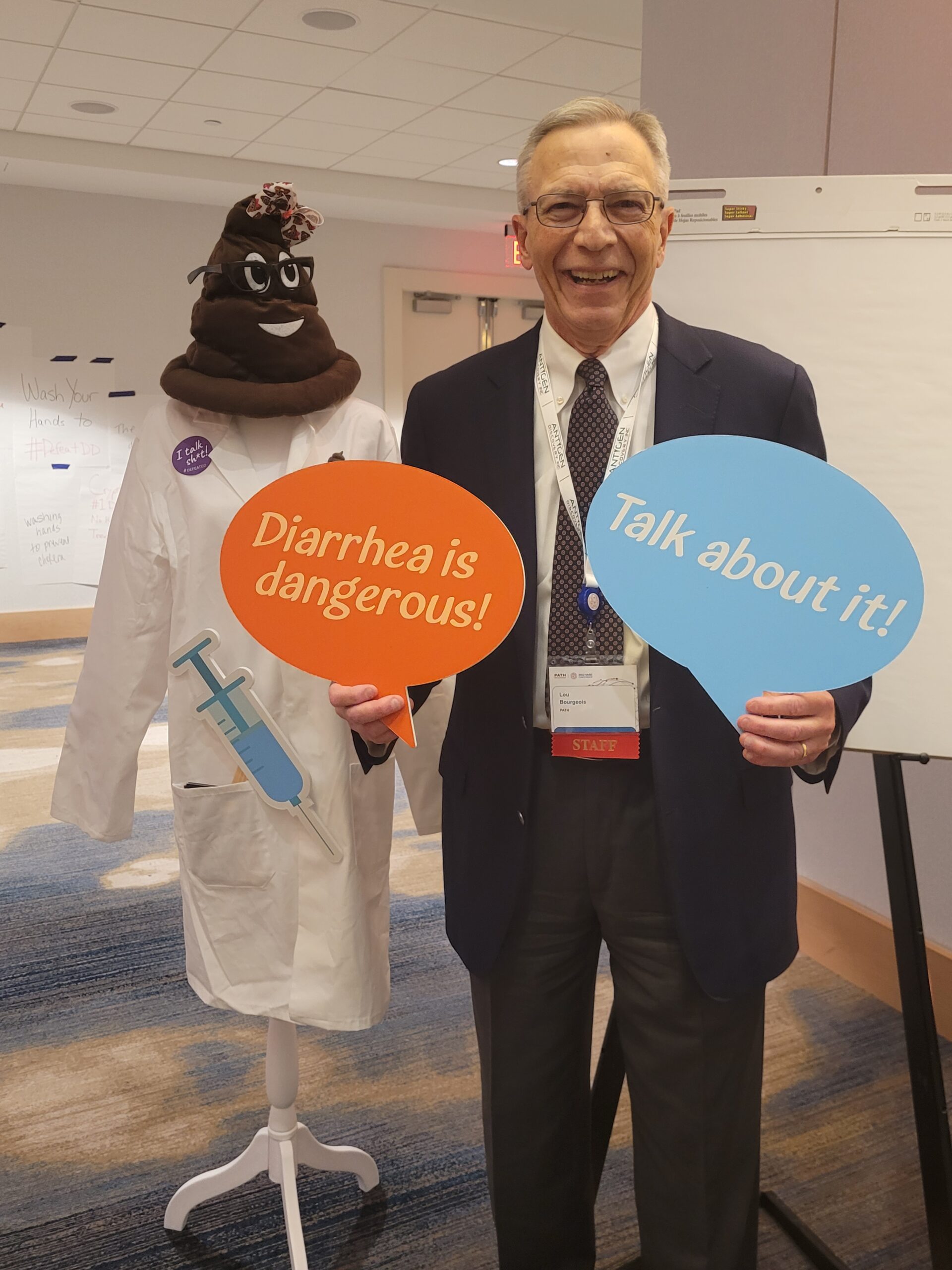 Lou stands next to a poo hat wearing a labcoat 