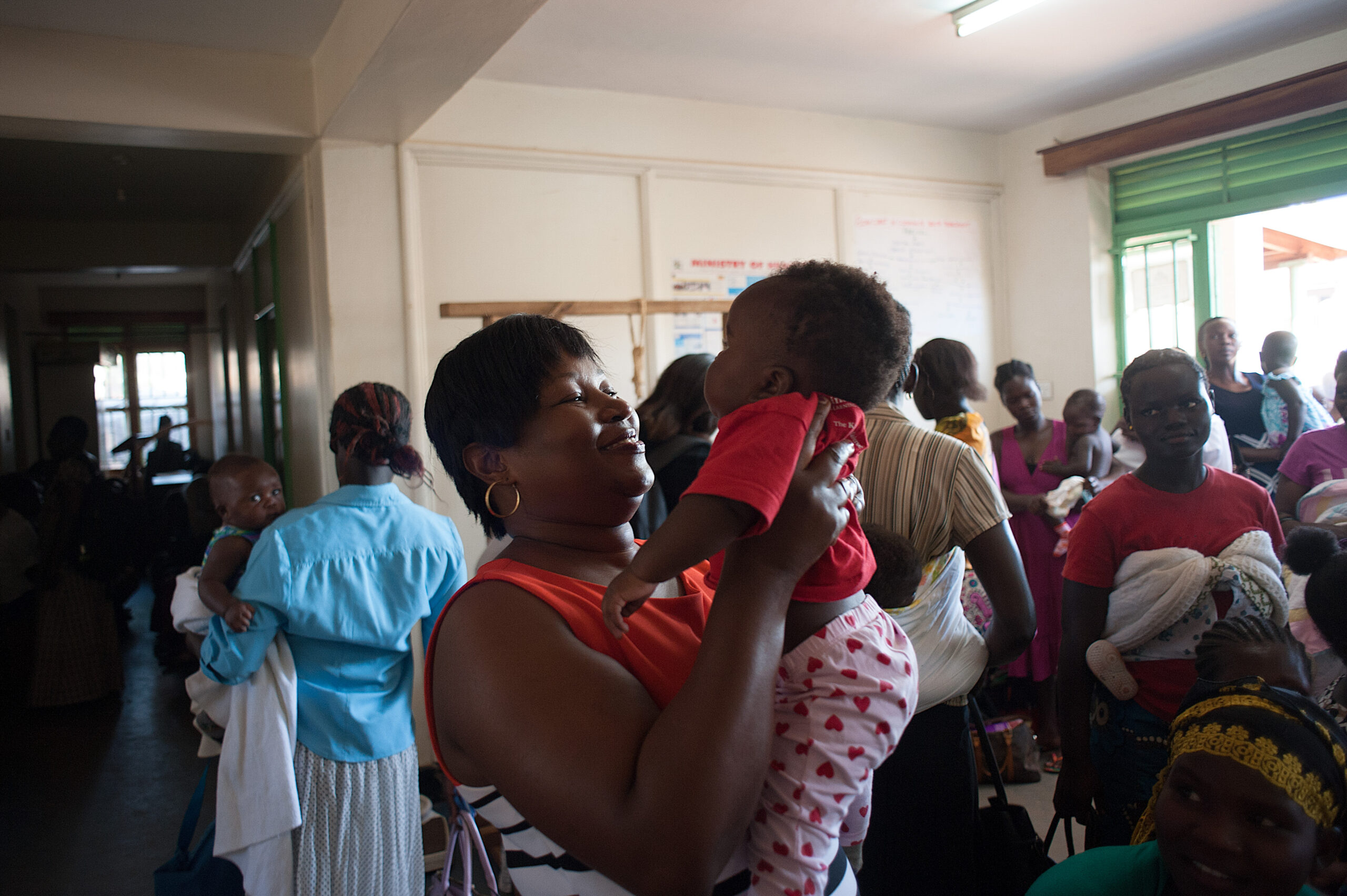 A woman in a striped top holds up a baby wearing a red shirt and polka dot pants inside a busy room. Other women and children are seen in the background, engaging in various activities.