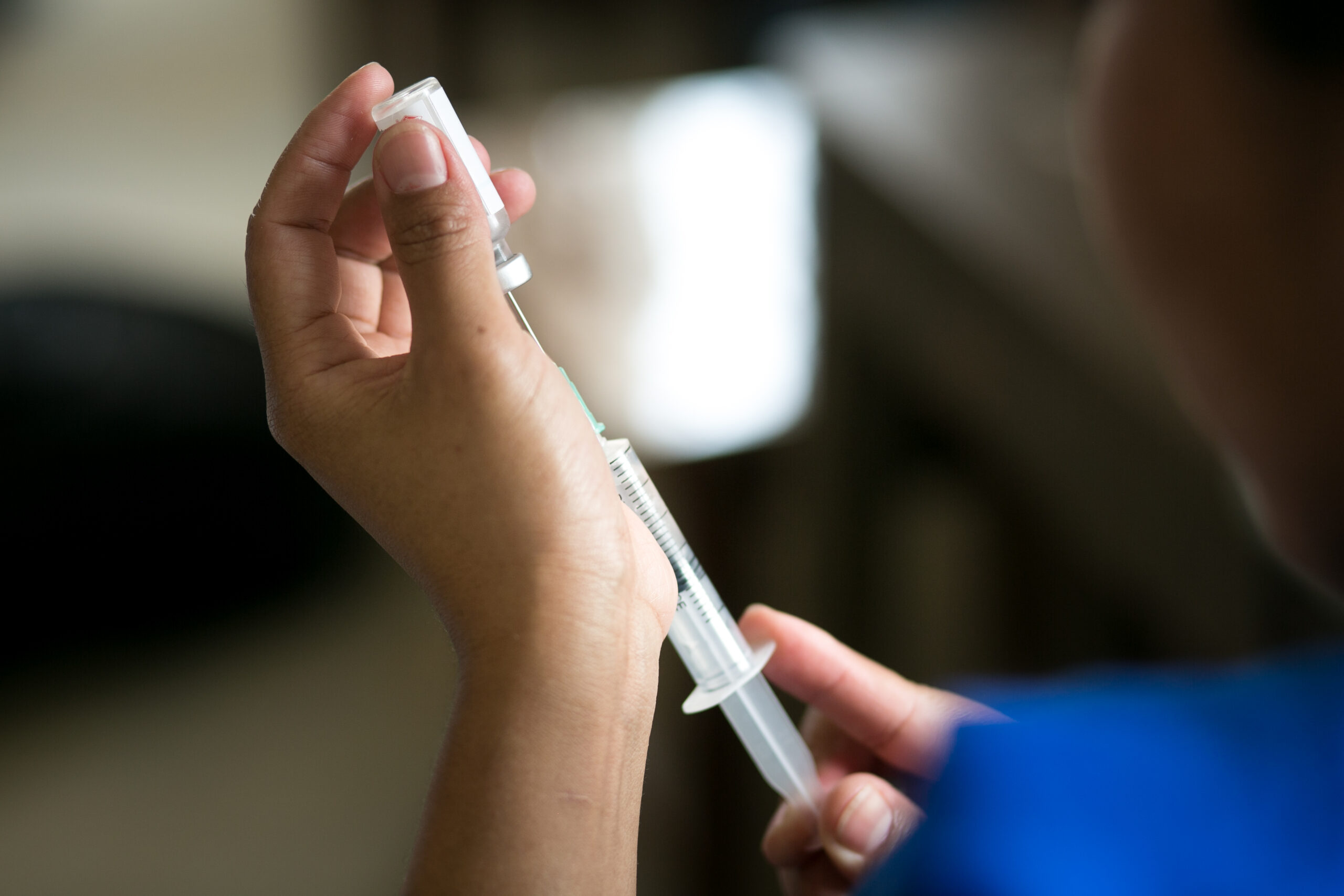 A person holding a syringe while preparing to administer an injection. The background is blurred, focusing on the hands and the syringe. The person is wearing a blue garment.