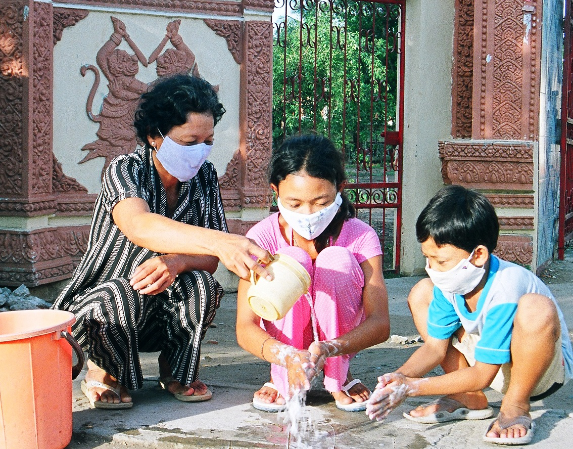 A woman in a striped outfit, along with a girl and a boy in colorful clothes, all wearing face masks, wash their hands with soapy water outdoors. They use a yellow container to pour water while kneeling next to a large orange bucket against a decorated wall.