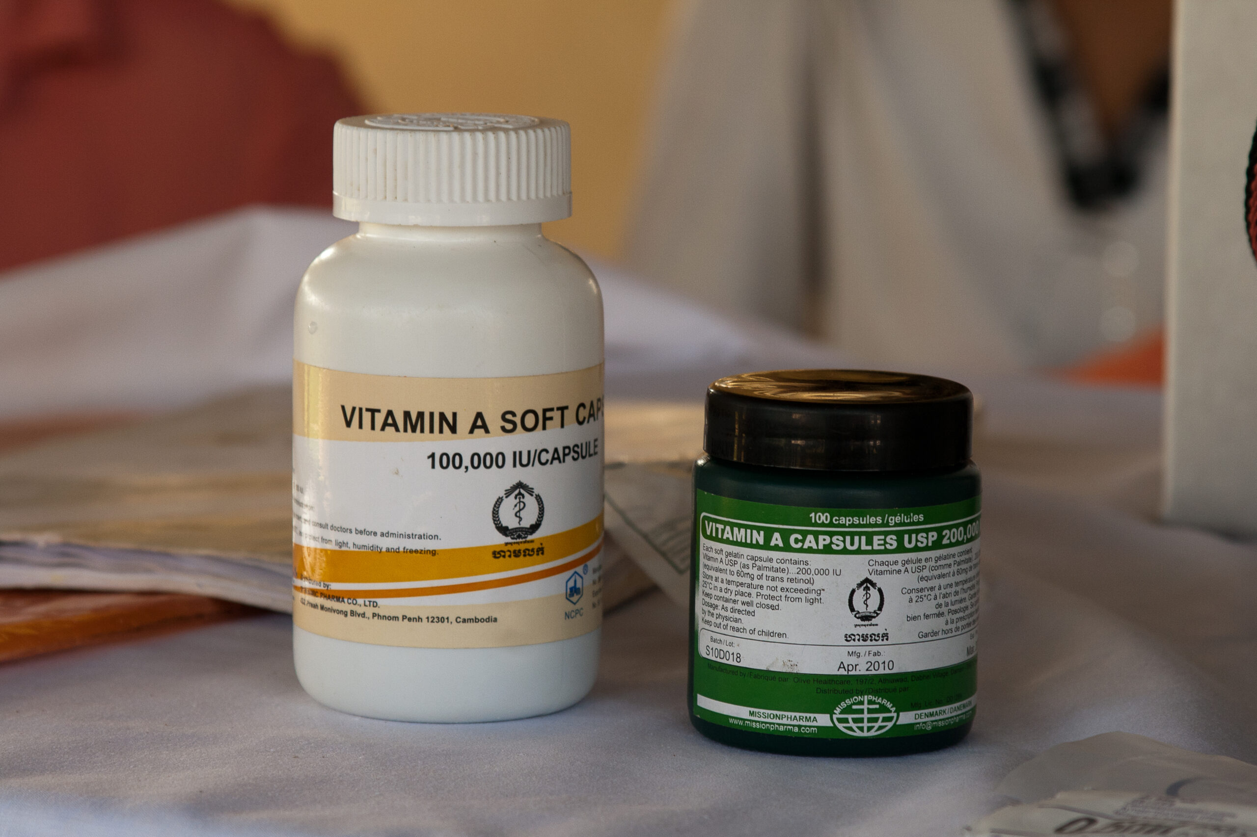 A bottle of Vitamin A soft capsules, labeled "100,000 IU/Capsule," stands next to a jar of Vitamin A capsules (USP 200,000 IU) on a table. The jar has a green label and indicates it contains 100 capsules, dated April 2013. Both containers are partially in view.