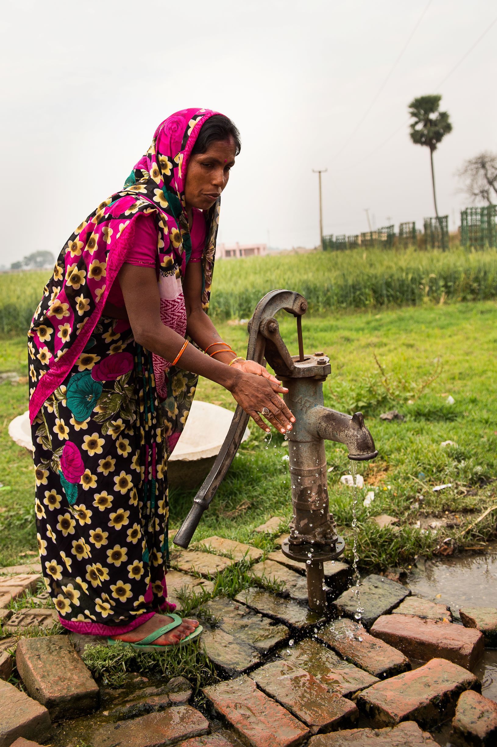 A woman in colorful traditional attire operates a hand pump in a rural setting. She is drawing water from the pump, with green grass and a few trees in the background. The ground around the pump is paved with bricks.