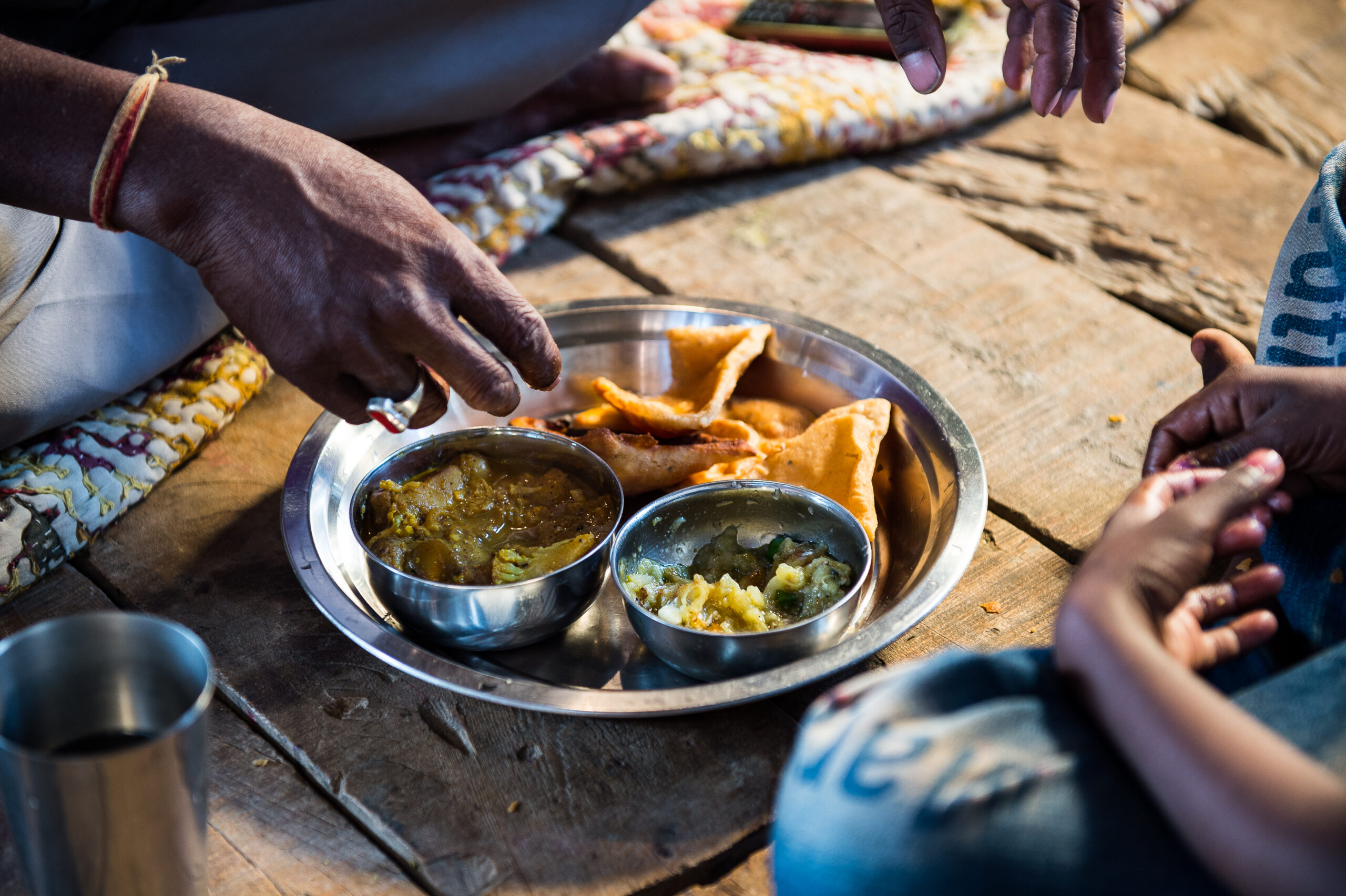 A close-up of people sitting on the floor, sharing a meal on a metal tray. The tray contains two bowls of curry and several fried pastries. One hand is reaching for food, and a child’s hands are also visible. The floor is wooden, and a patterned cloth is seen underneath.