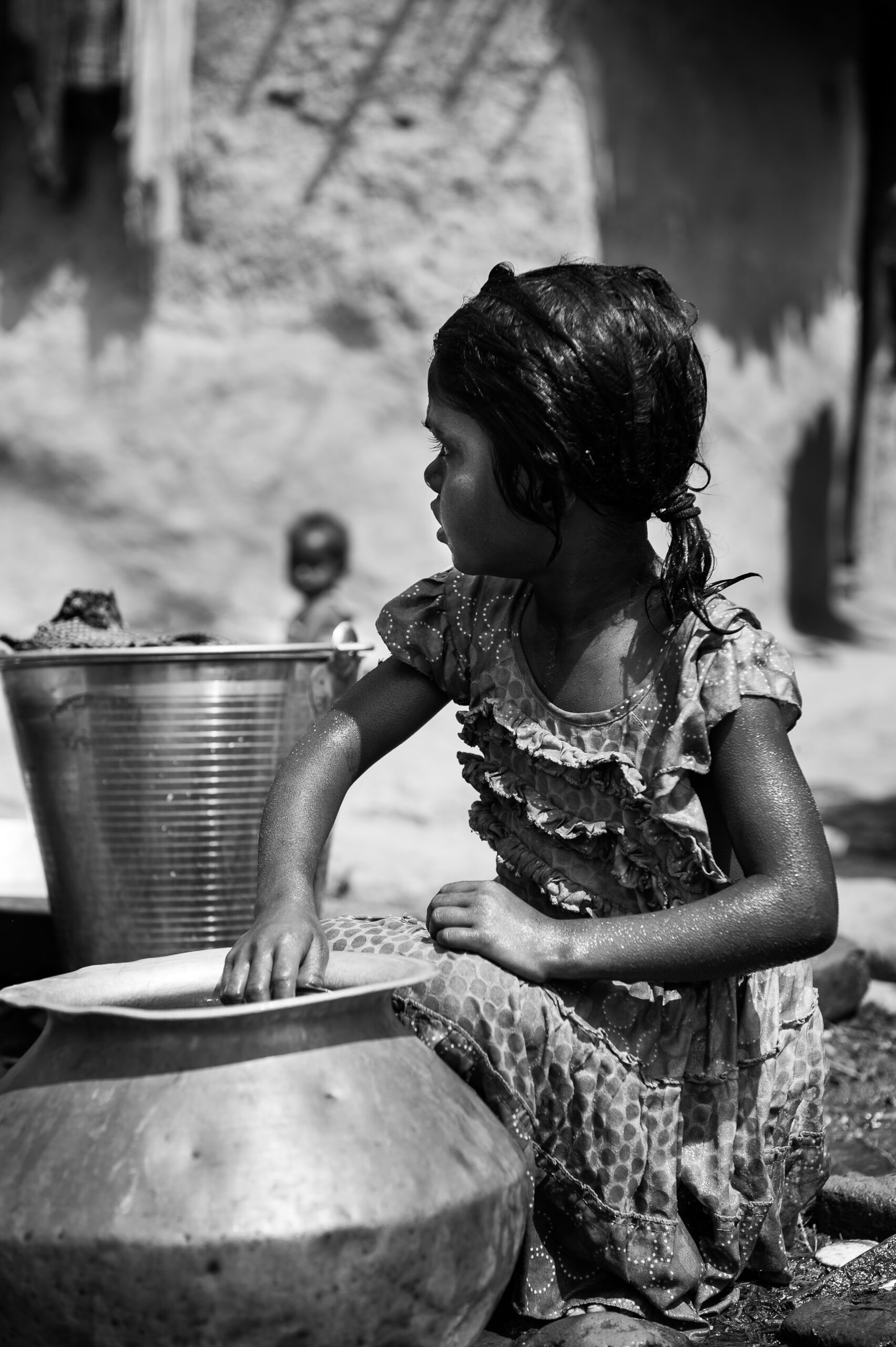 A young girl in a patterned dress sits outdoors, washing a large metal pot while looking to the side. Another child is in the blurred background beside a large metal basin. The setting appears to be a modest, sunlit rural environment. Image is in black and white.