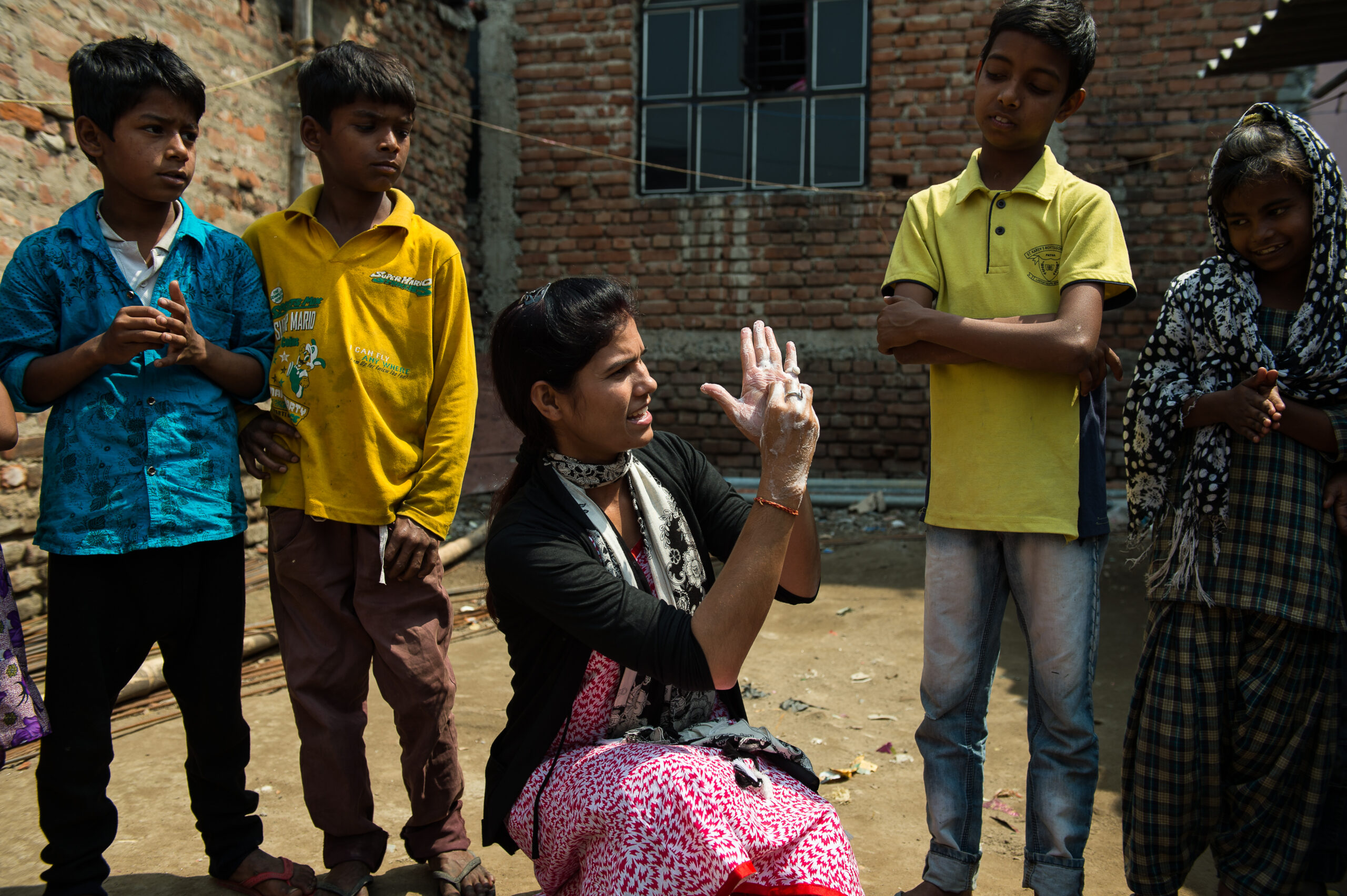 A woman kneels, demonstrating hand gestures to a group of five children. They are standing outside in a sunny area with brick buildings in the background. The children watch attentively, dressed in bright, casual clothing.