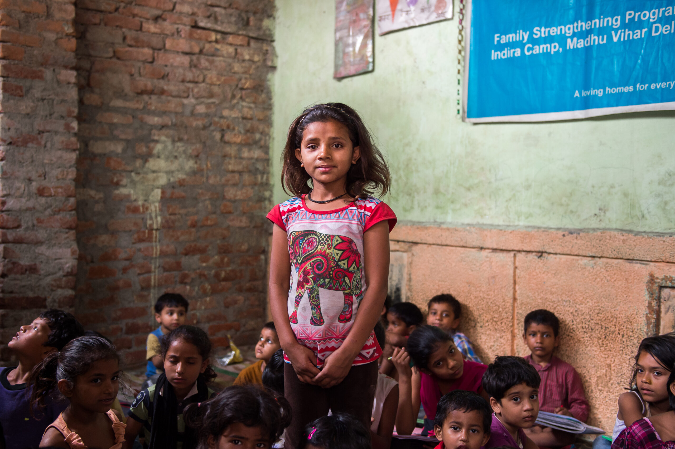 A young girl in a colorful shirt stands confidently in the middle of a brick-walled classroom, surrounded by other children seated on the floor. A blue banner hangs in the background, partially visible. The environment appears educational and community-focused.