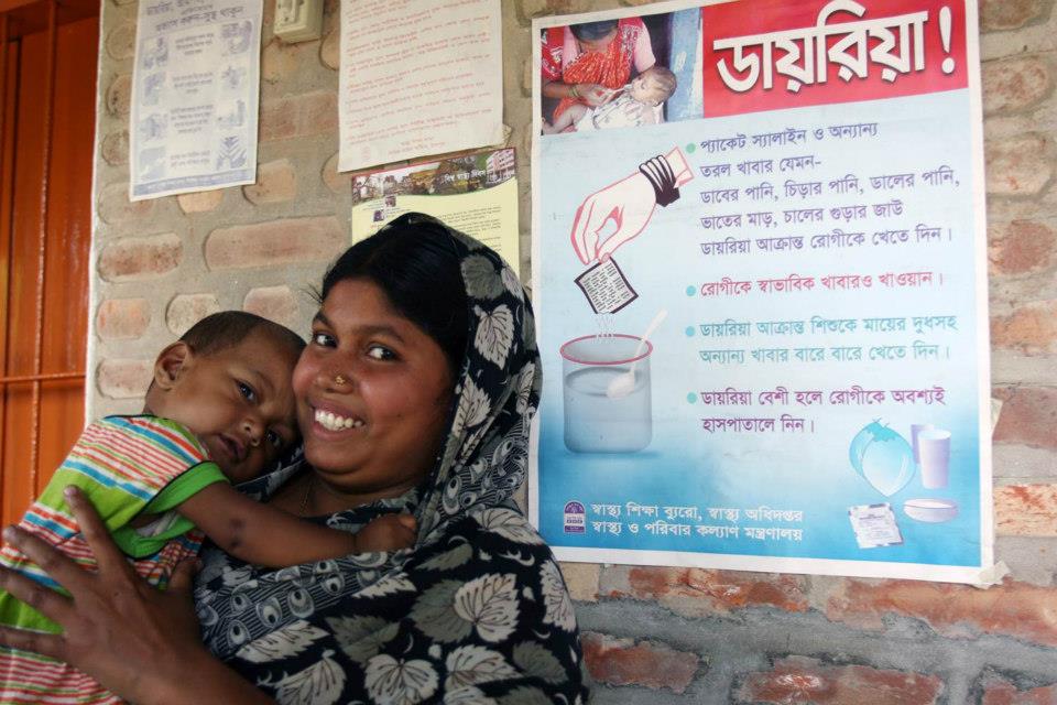 A woman smiles at the camera while holding a child in front of a brick wall. A poster with Bengali text about diarrhea prevention and treatment is visible behind them, showing illustrations of clean water, medicines, and steps for ensuring hygiene and health.