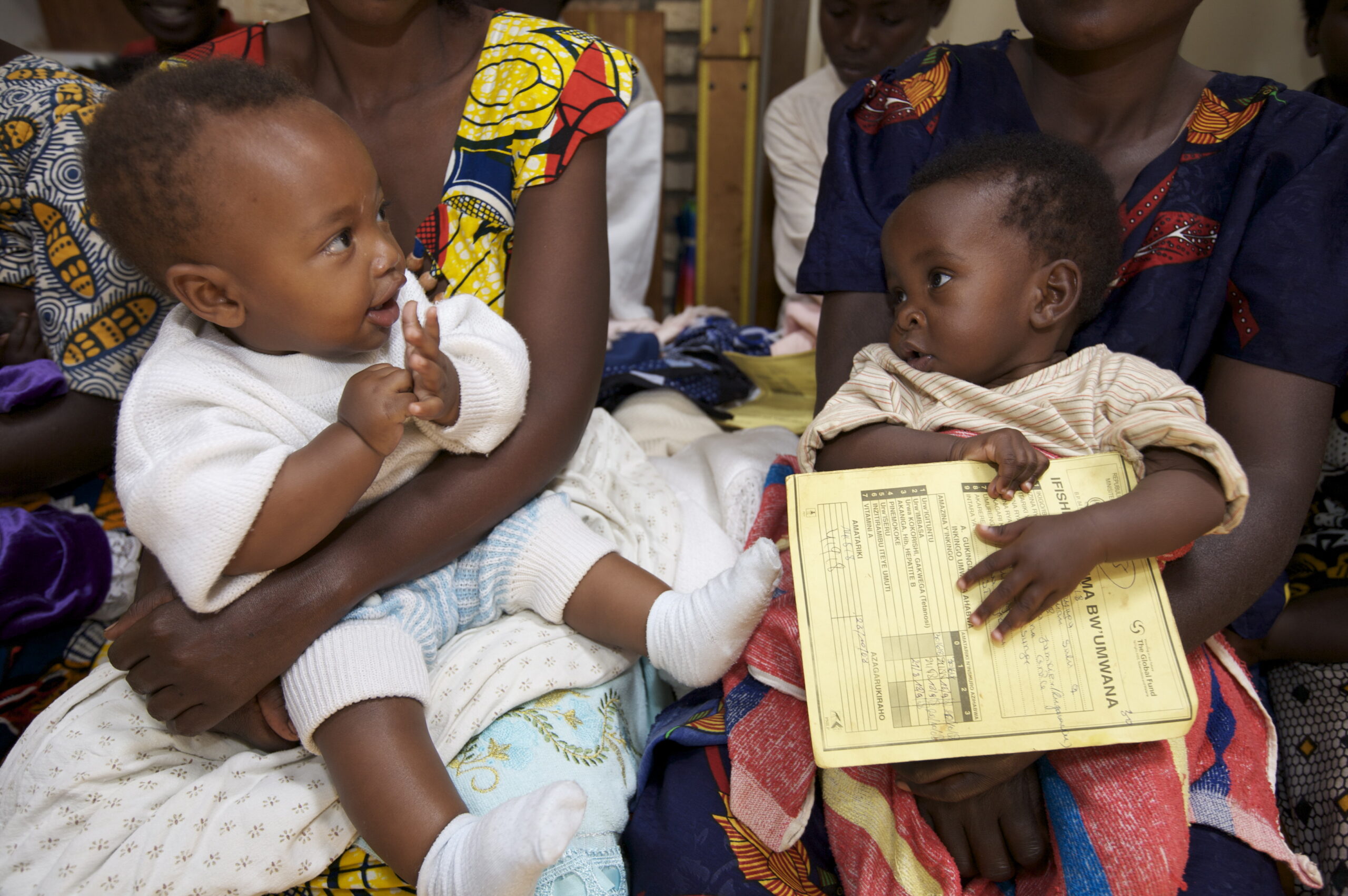 Two babies are sitting on the laps of adults. One baby wears a white outfit, looking inquisitively at the other, who holds a document. The adults wear colorful patterned clothing, seated closely together.