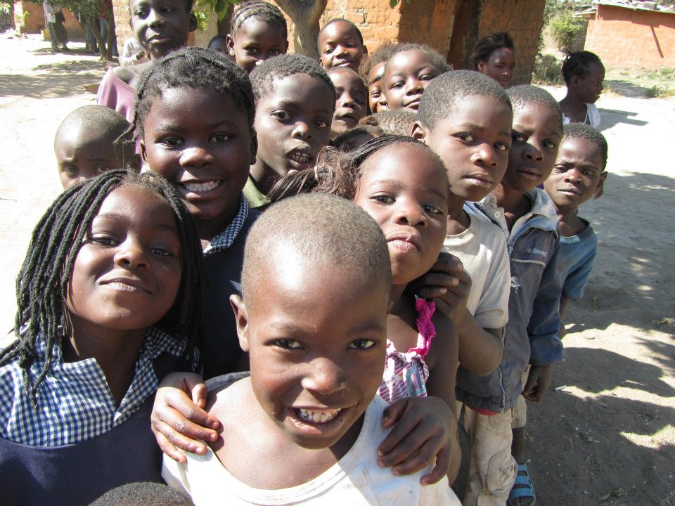 A group of smiling children, most wearing casual clothes, stand closely together in an outdoor setting. The children are diverse in appearance and are lined up, some with hands on each other's shoulders. The background includes trees and a building.