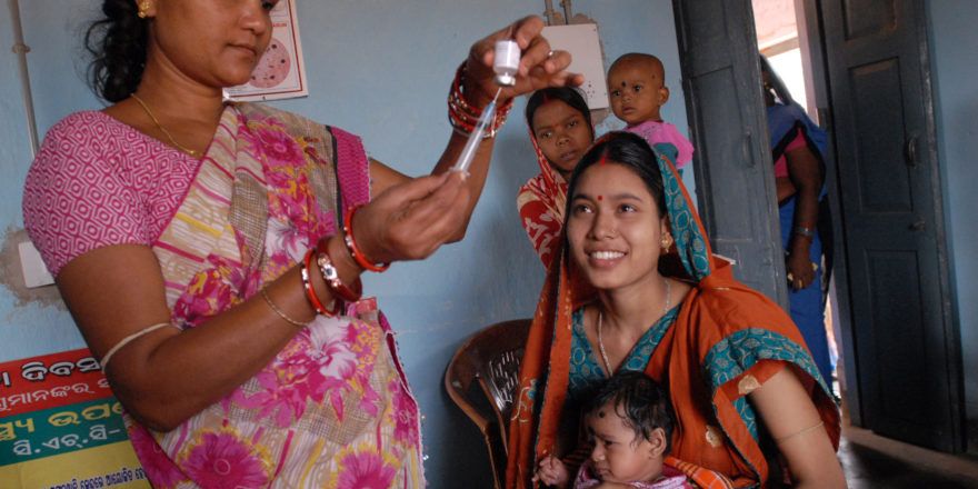 A healthcare worker in a pink sari is preparing a syringe while a woman in an orange sari, holding a baby, sits smiling. Another woman holding a child stands in the background near an open door. The setting appears to be a clinic with posters on the wall.