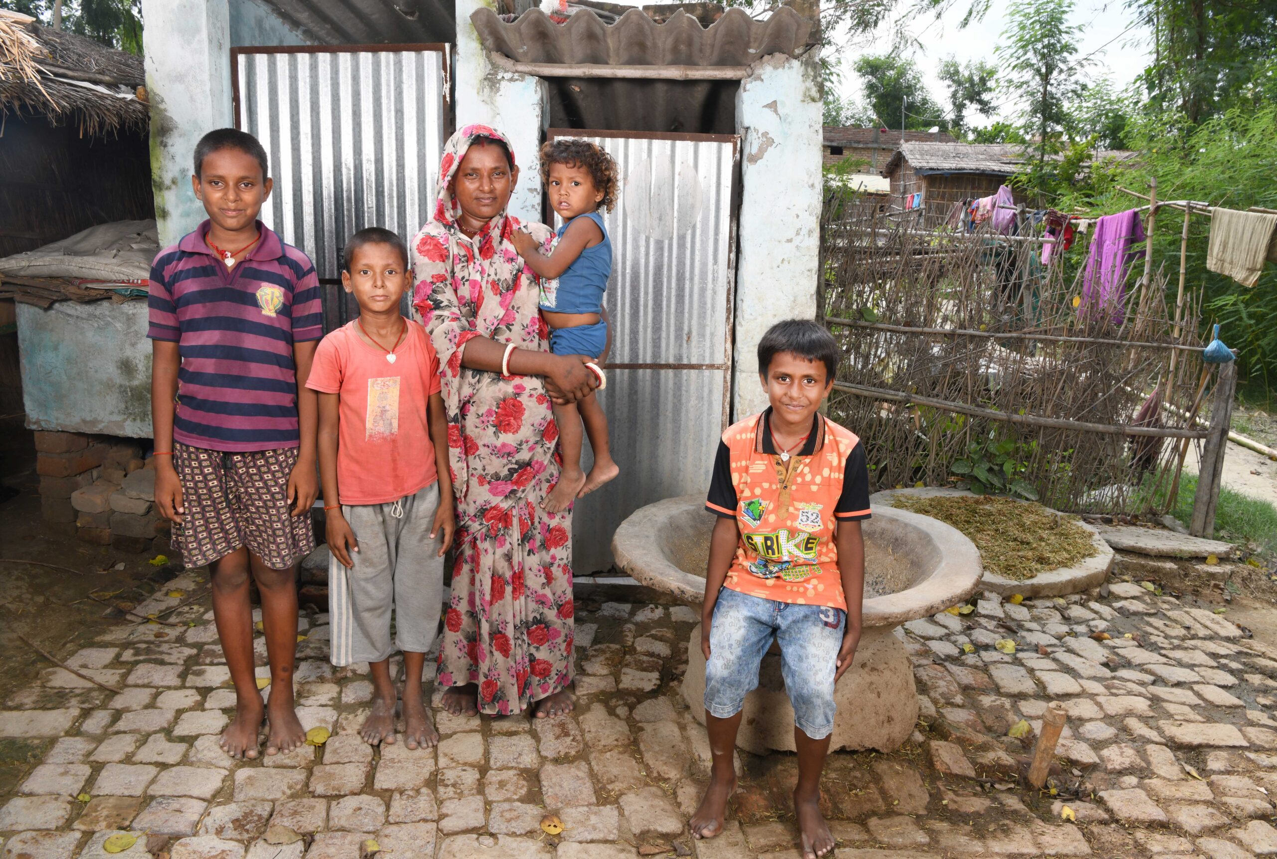 A group of five people, including a woman holding a young child, stand on a cobblestone path in front of a small building. They are outdoors, surrounded by greenery and a laundry line with clothes.