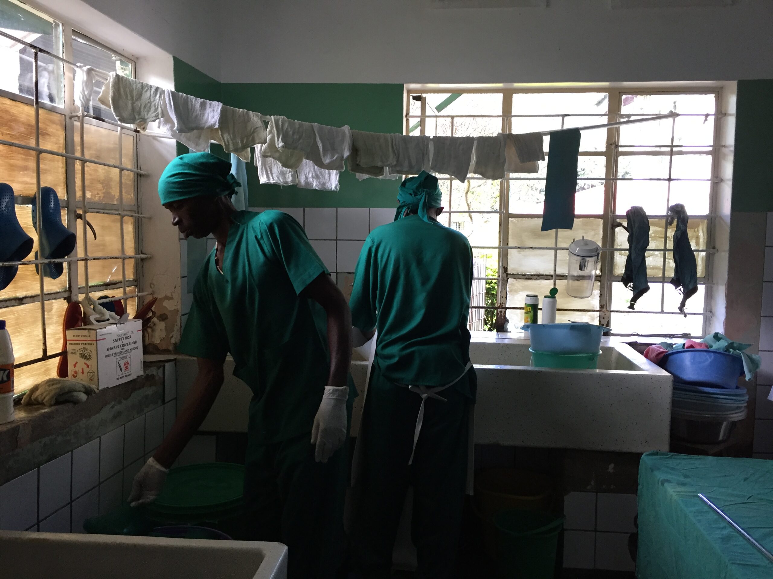 Two healthcare workers in green scrubs are seen working in a dimly lit medical room. One is at a counter, and the other is near a sink. Surgical towels hang on a line above. Various medical supplies are on the counter, with sunlight filtering through the windows.