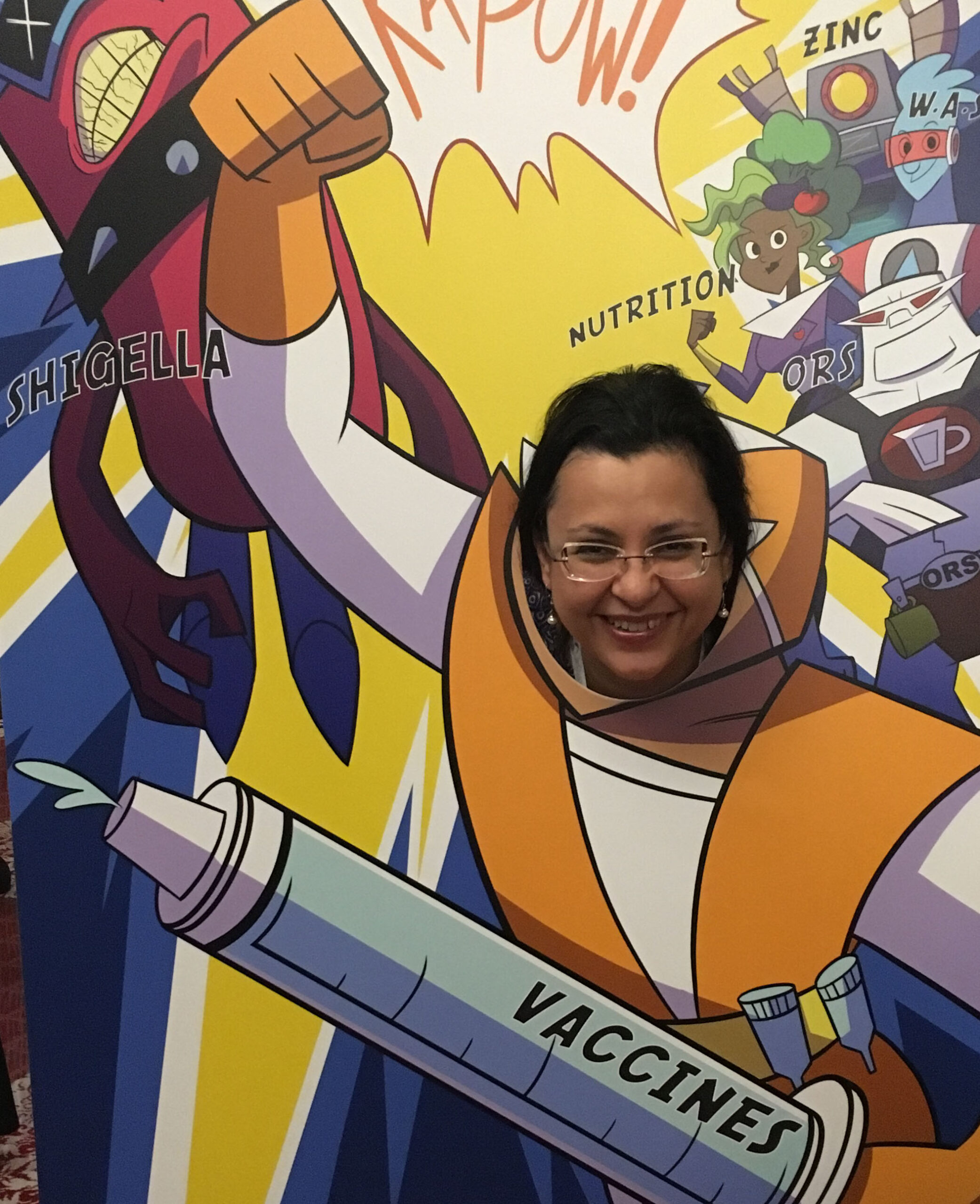 A woman playfully poses behind a vibrant cartoon cutout featuring a superhero with a large syringe labeled "Vaccines". The background includes colorful elements and words like "Shigella," "Nutrition," and "Zinc.